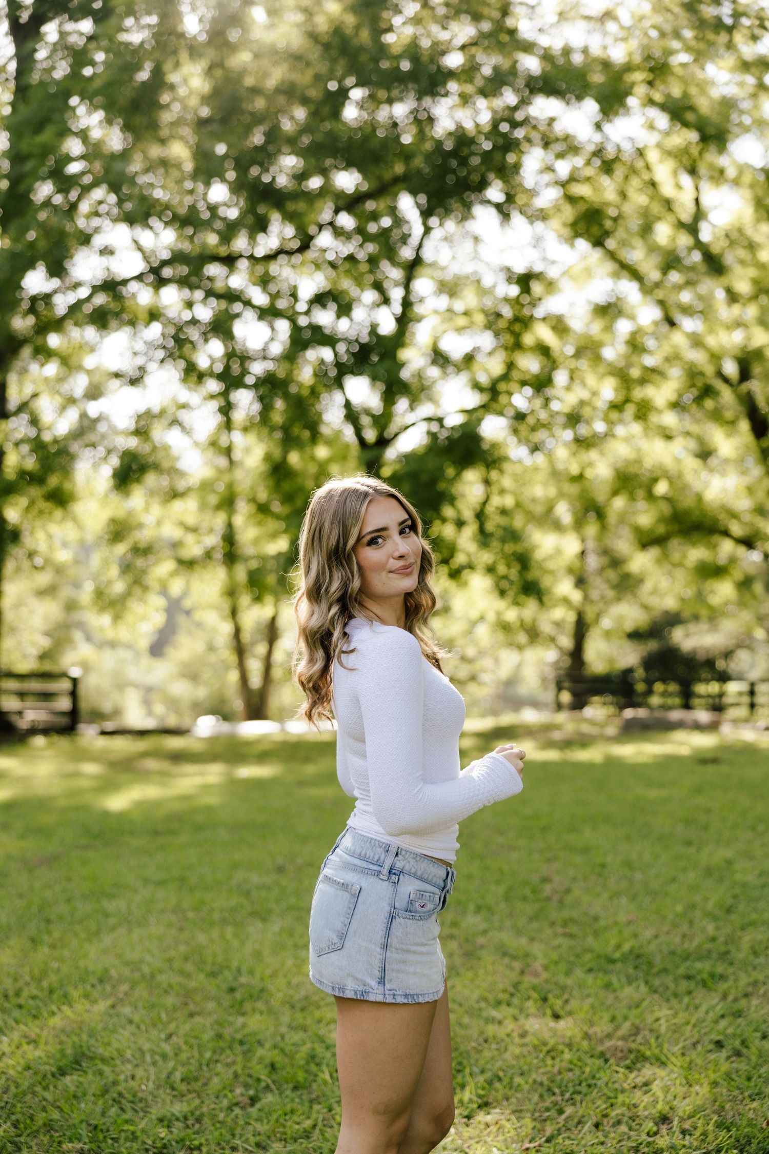 Young woman in white long-sleeve top and denim shorts smiling in a lush green park on a sunny day.