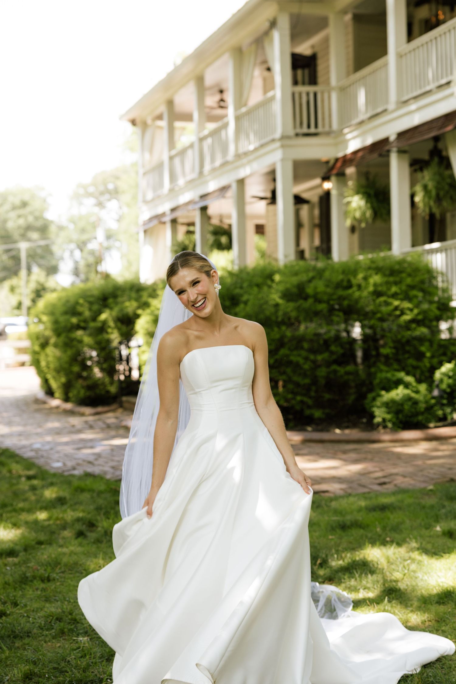 Smiling bride in strapless white ball gown with veil poses outdoors in front of elegant two-story white building.