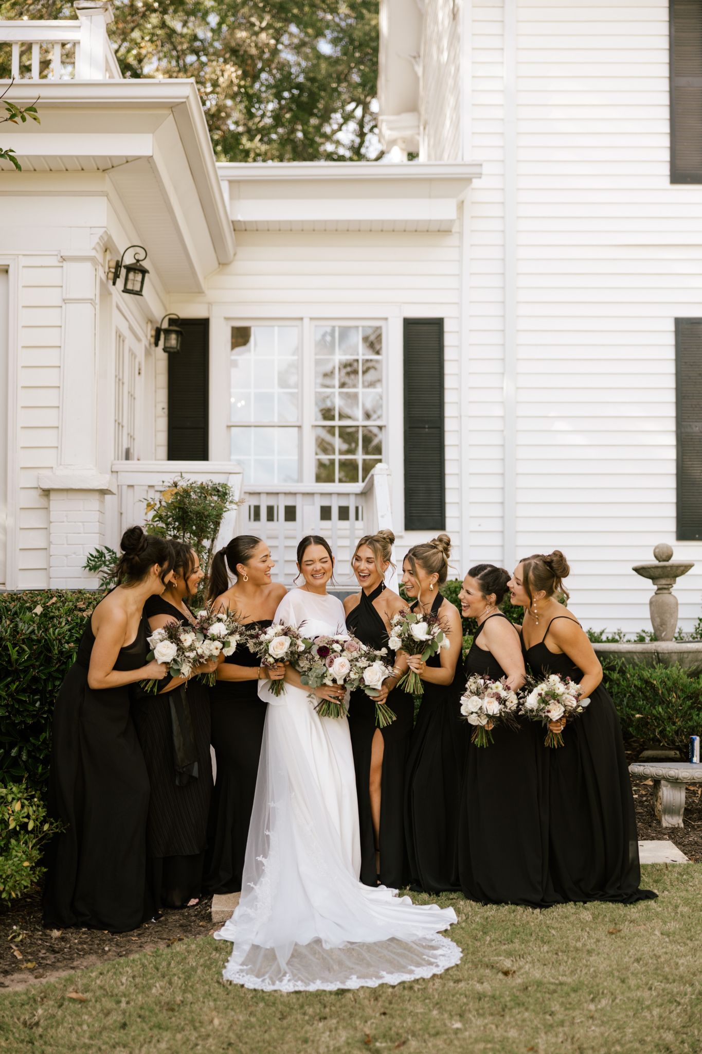 Bride in white gown poses with bridesmaids in black dresses outside a white colonial-style home.