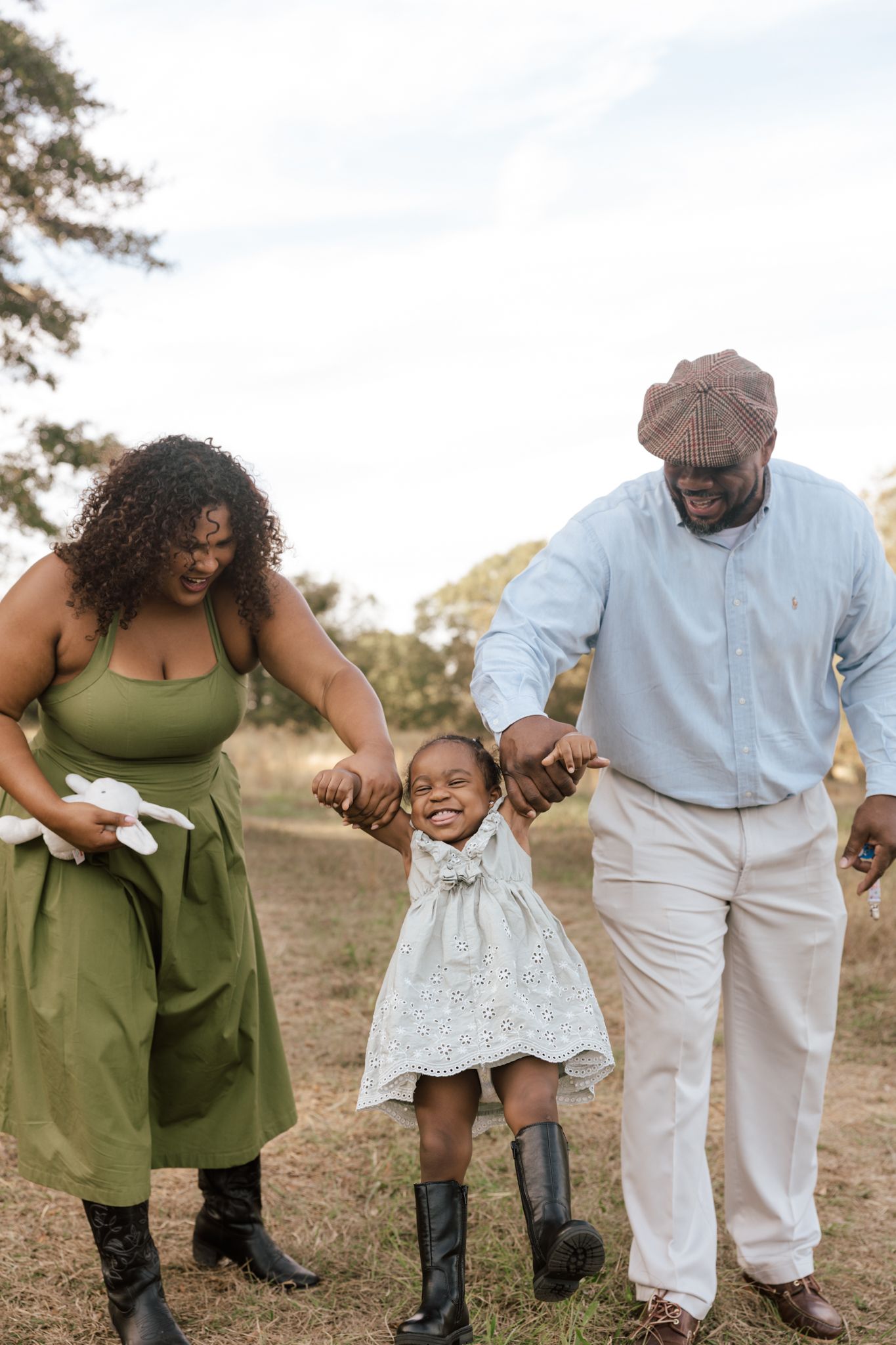 A joyful Black family with parents holding hands of their smiling little girl in a white dress outdoors.