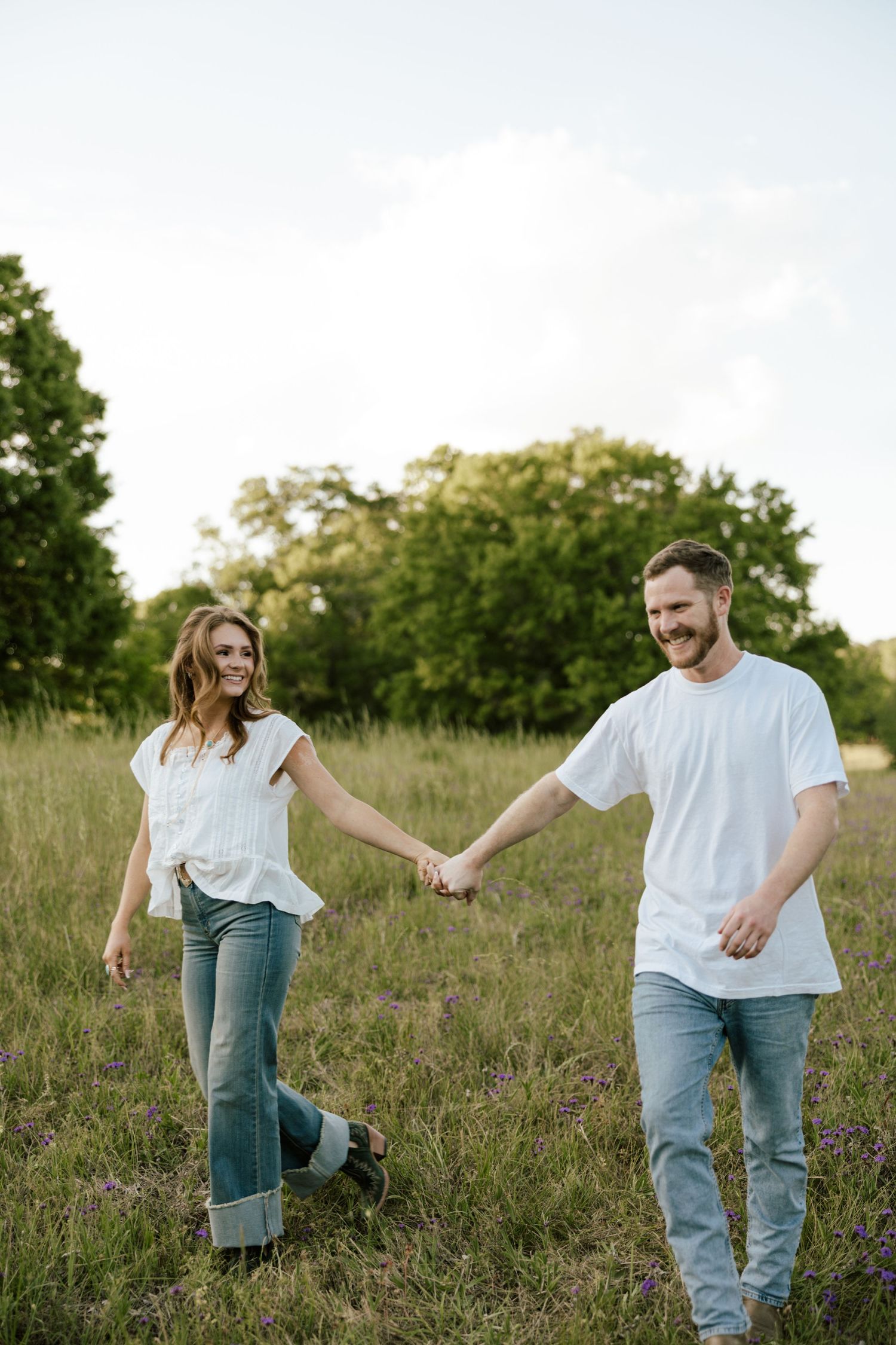 A smiling couple holds hands while walking through a lush green field, both wearing white tops and jeans.