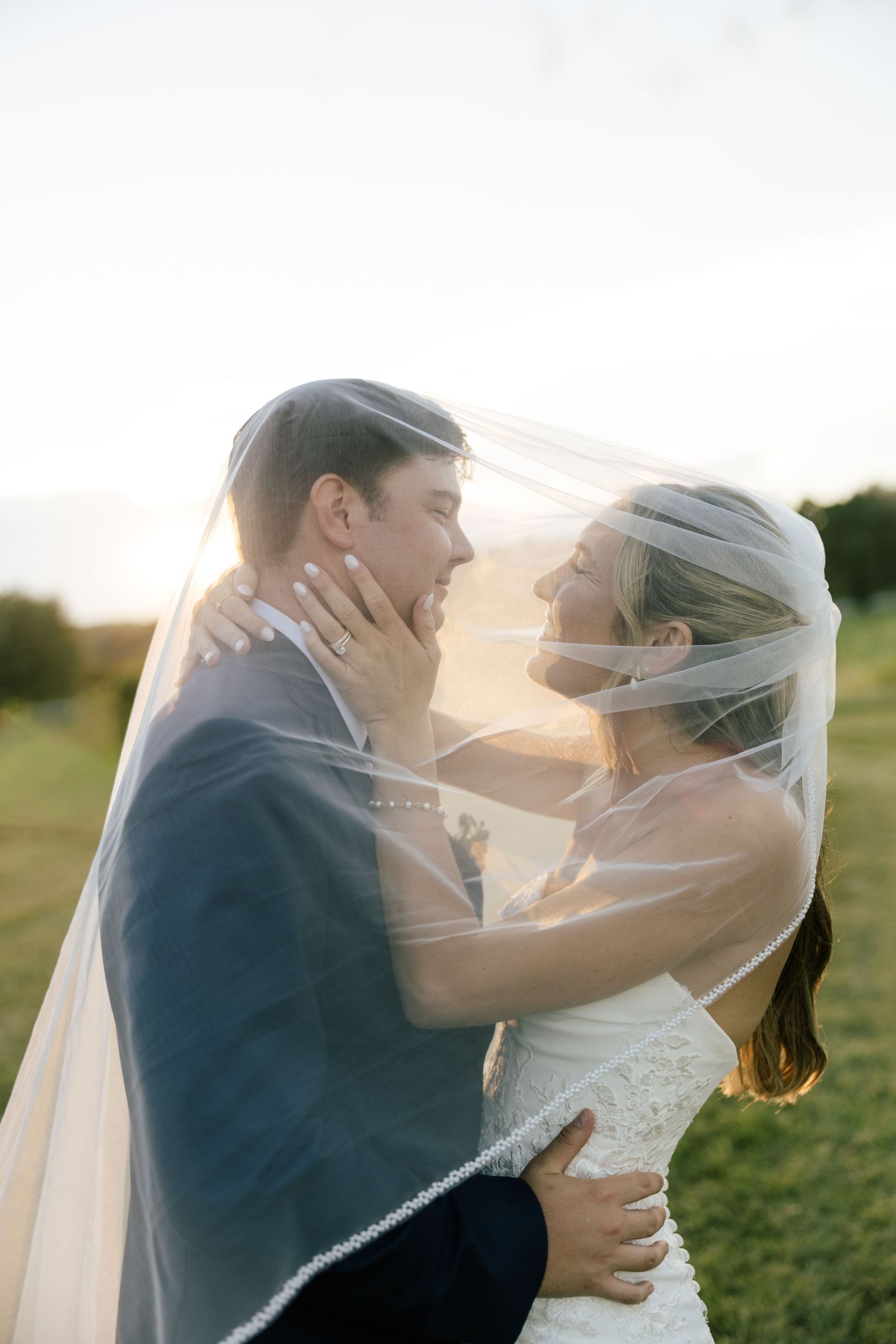 Bride and groom share romantic moment under wedding veil at golden hour in a green field.