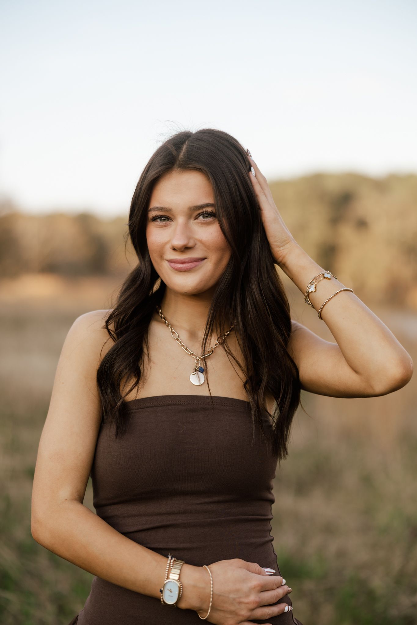 Young woman in brown tube top with gold jewelry smiles outdoors in a field during golden hour.