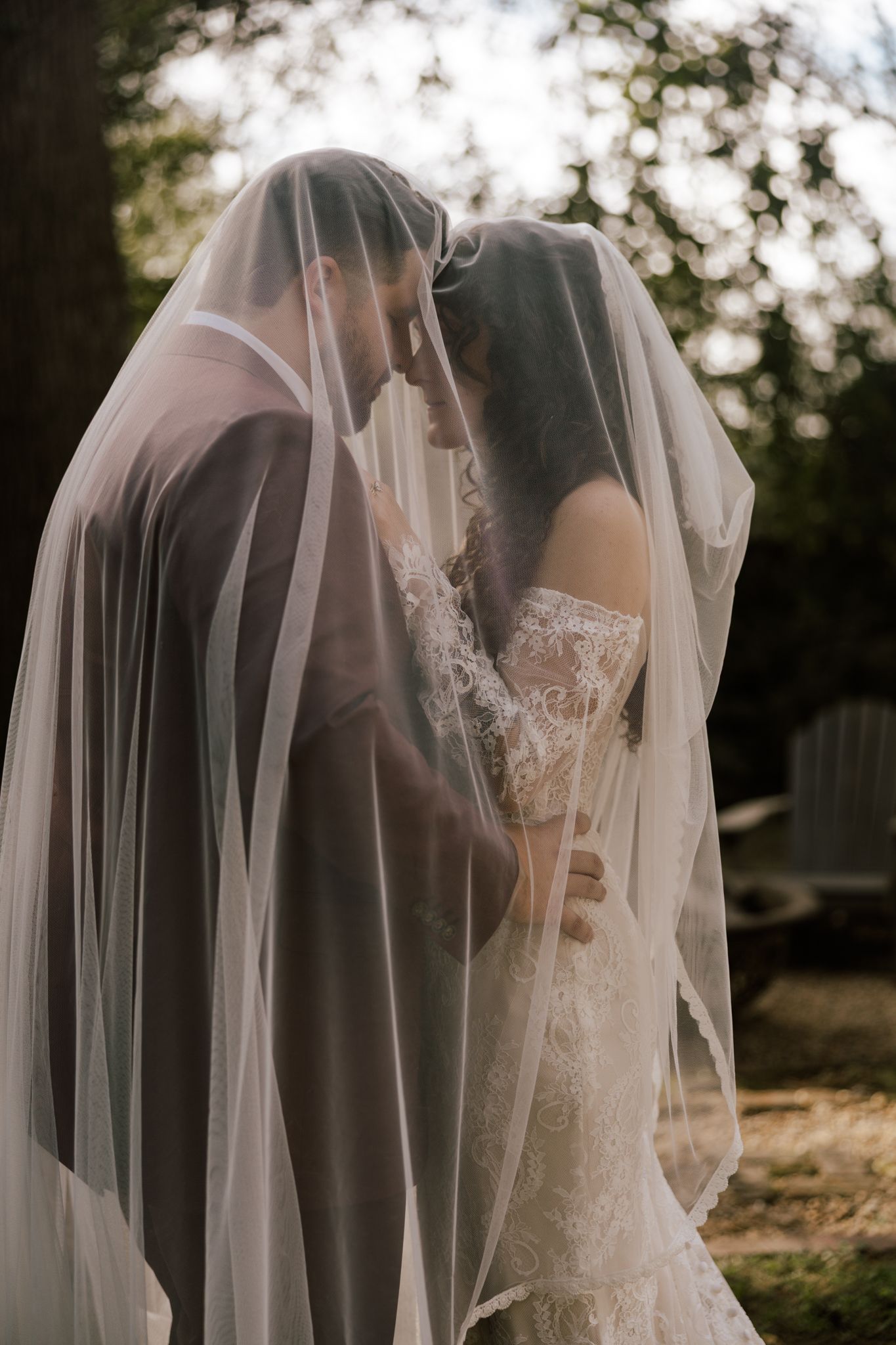 Bride and groom share an intimate moment under a flowing white veil, wearing lace wedding gown outdoors.