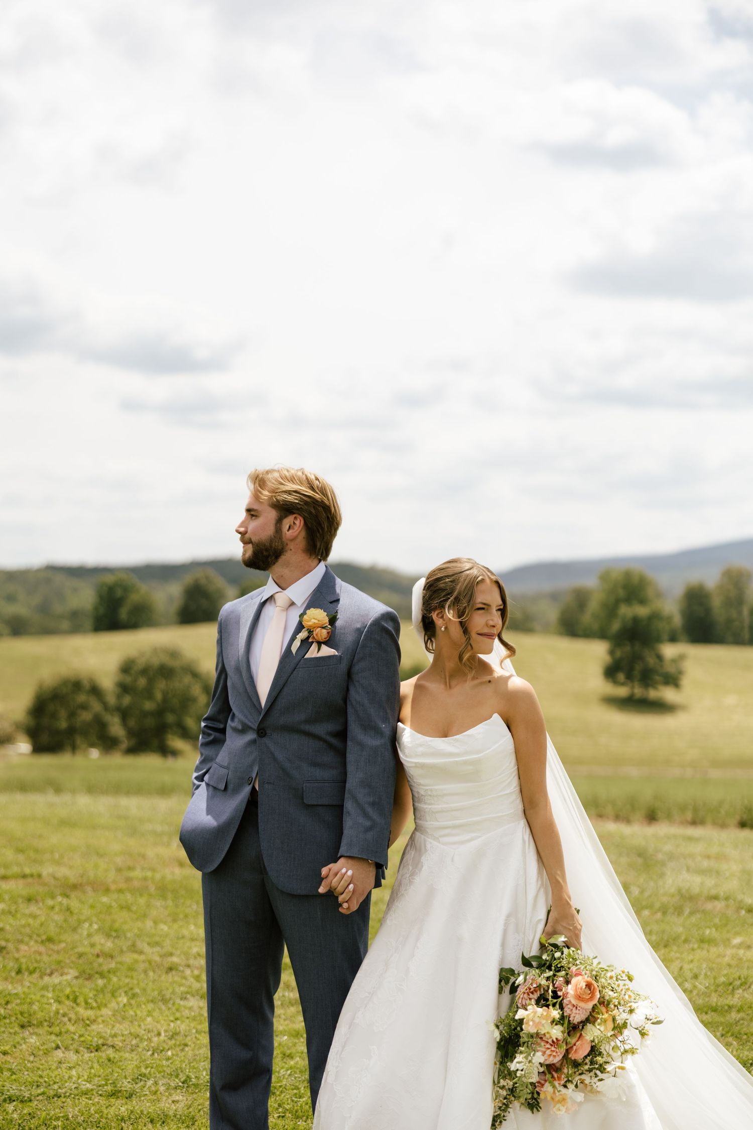 A bride and groom hold hands in a scenic green field, she in a white gown with bouquet, he in a navy suit.