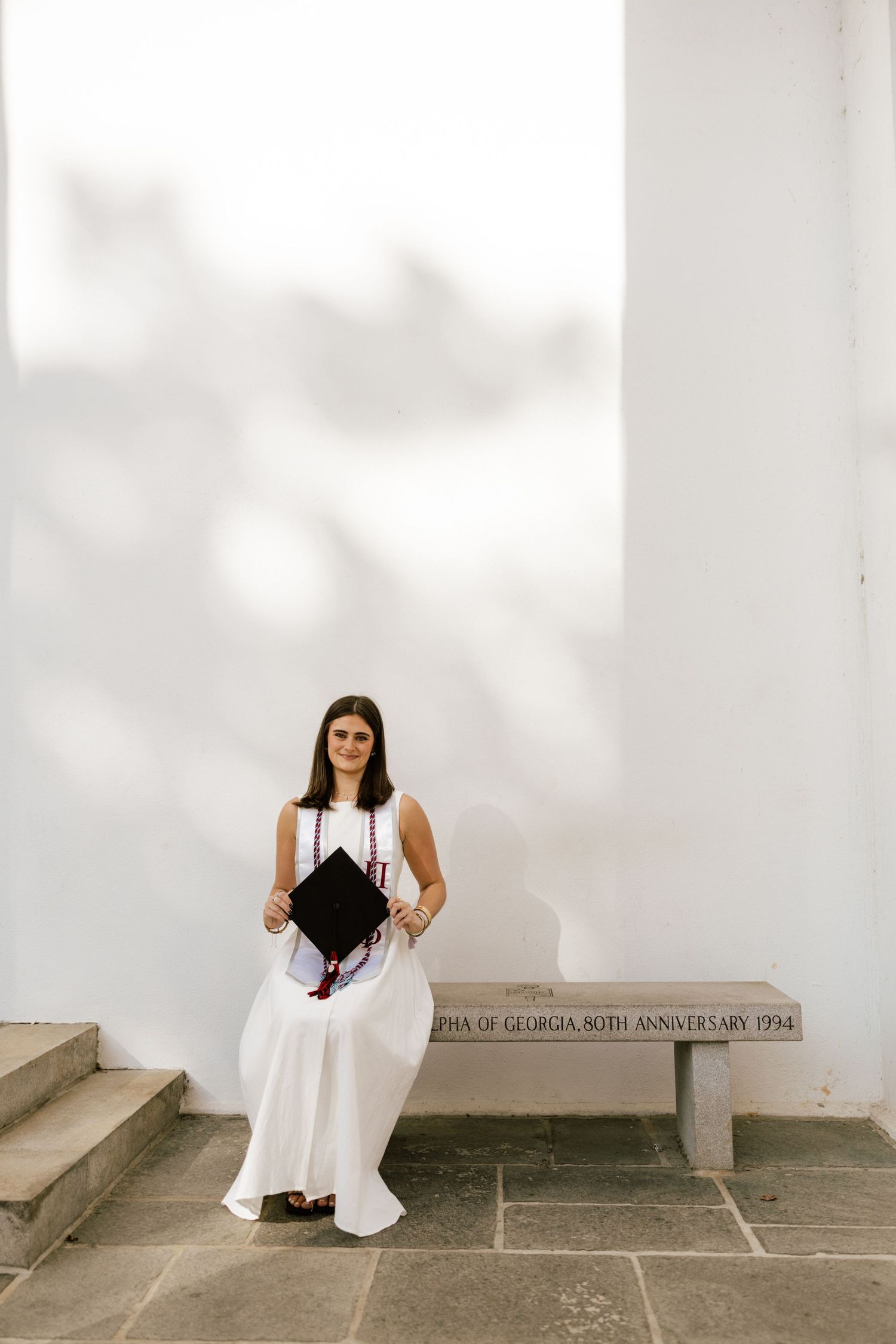 Young woman in white dress holding graduation cap, seated on anniversary bench at University of Georgia.
