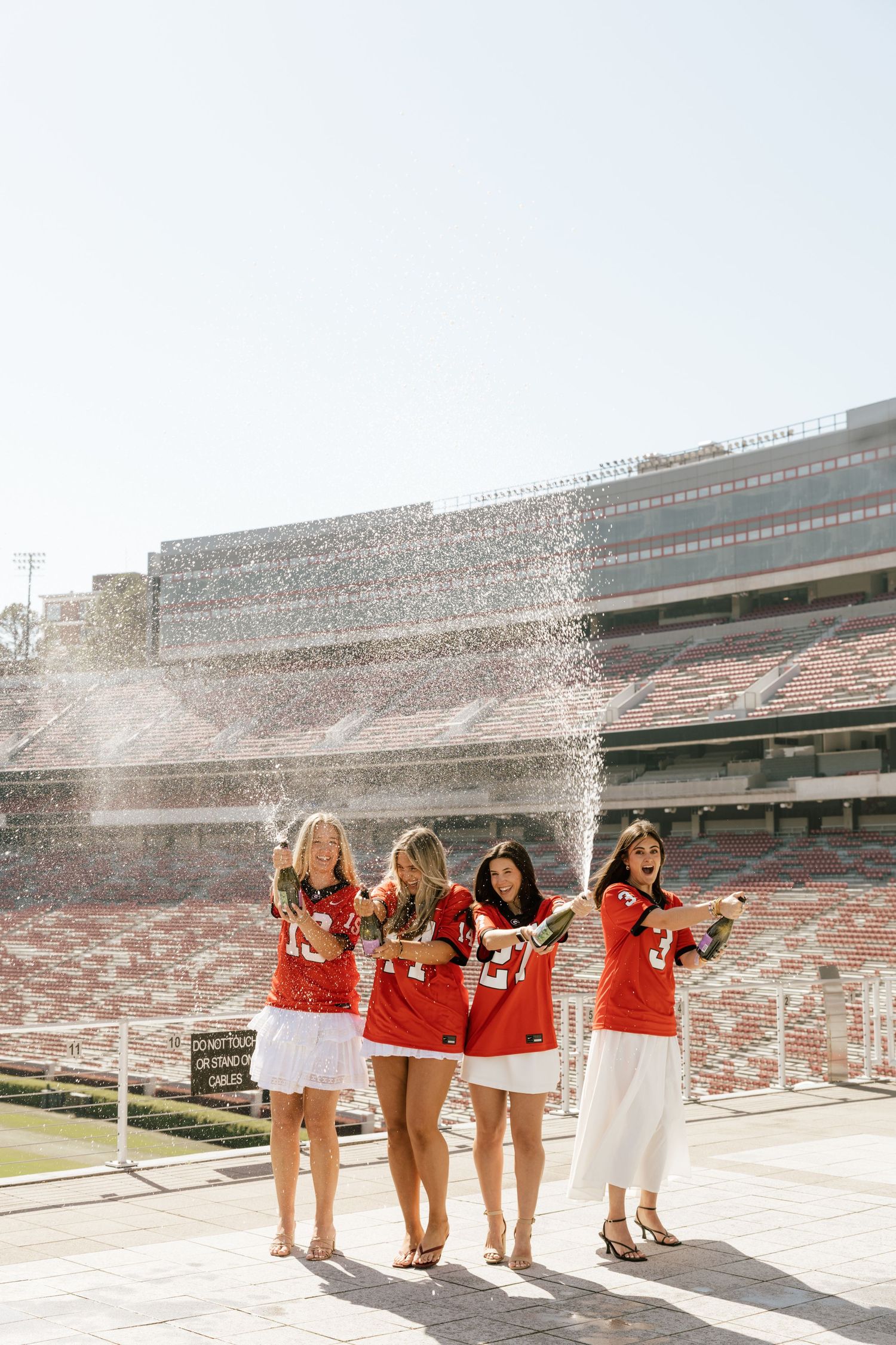 Four women in red jerseys celebrate with champagne in an empty stadium on a sunny day.