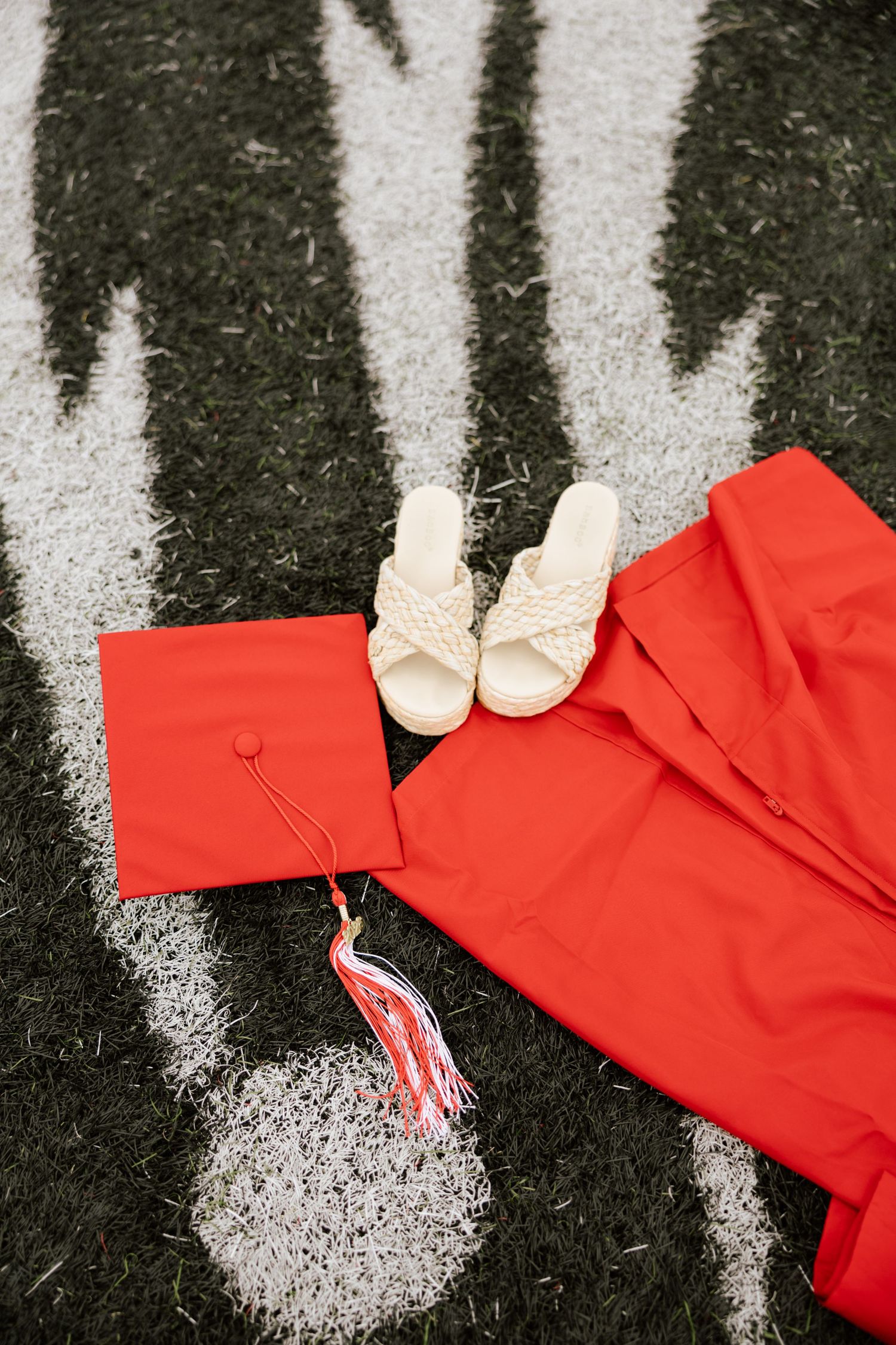 Red graduation cap, gown, and white sandals laid on green turf field with white yard line markings.