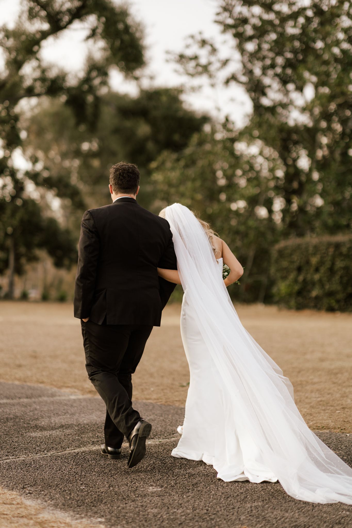 A bride and groom walk hand in hand away from the camera, surrounded by lush green trees on their wedding day.