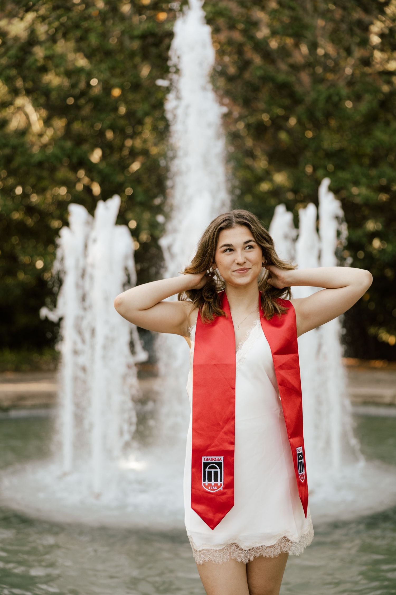 A smiling graduate poses in a white dress and red graduation stole in front of a fountain surrounded by lush greenery.