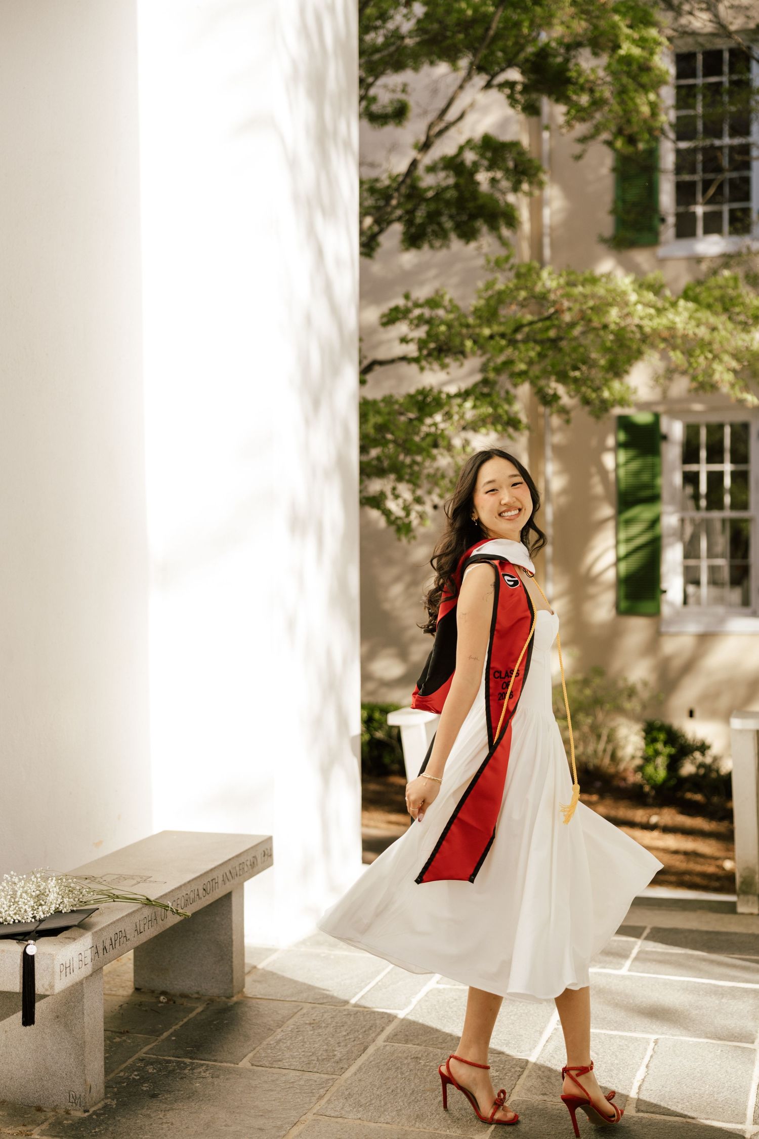 Young woman in white dress and red graduation sash smiles outdoors near a historic building with green trees.