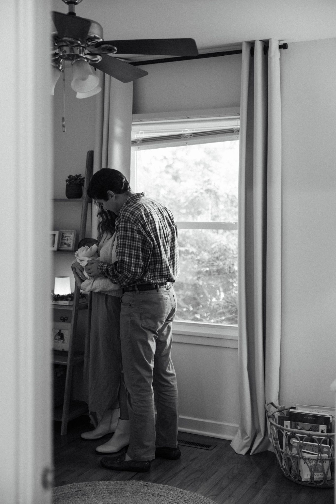 Black and white photo of a couple sharing a tender moment by a window in a cozy room with ceiling fan.