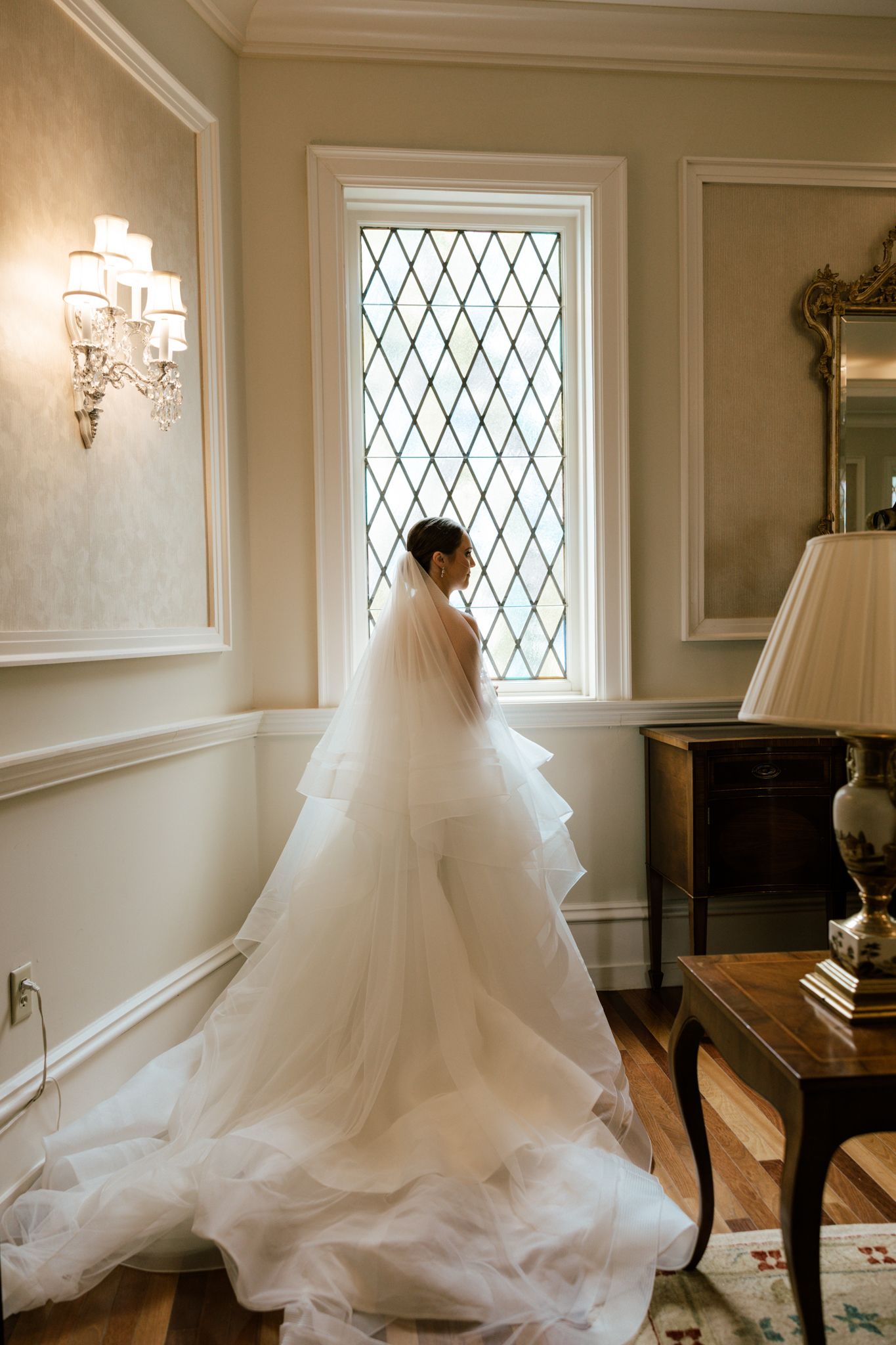 An elegant wedding dress with a flowing train displayed in a room with diamond-paned windows.