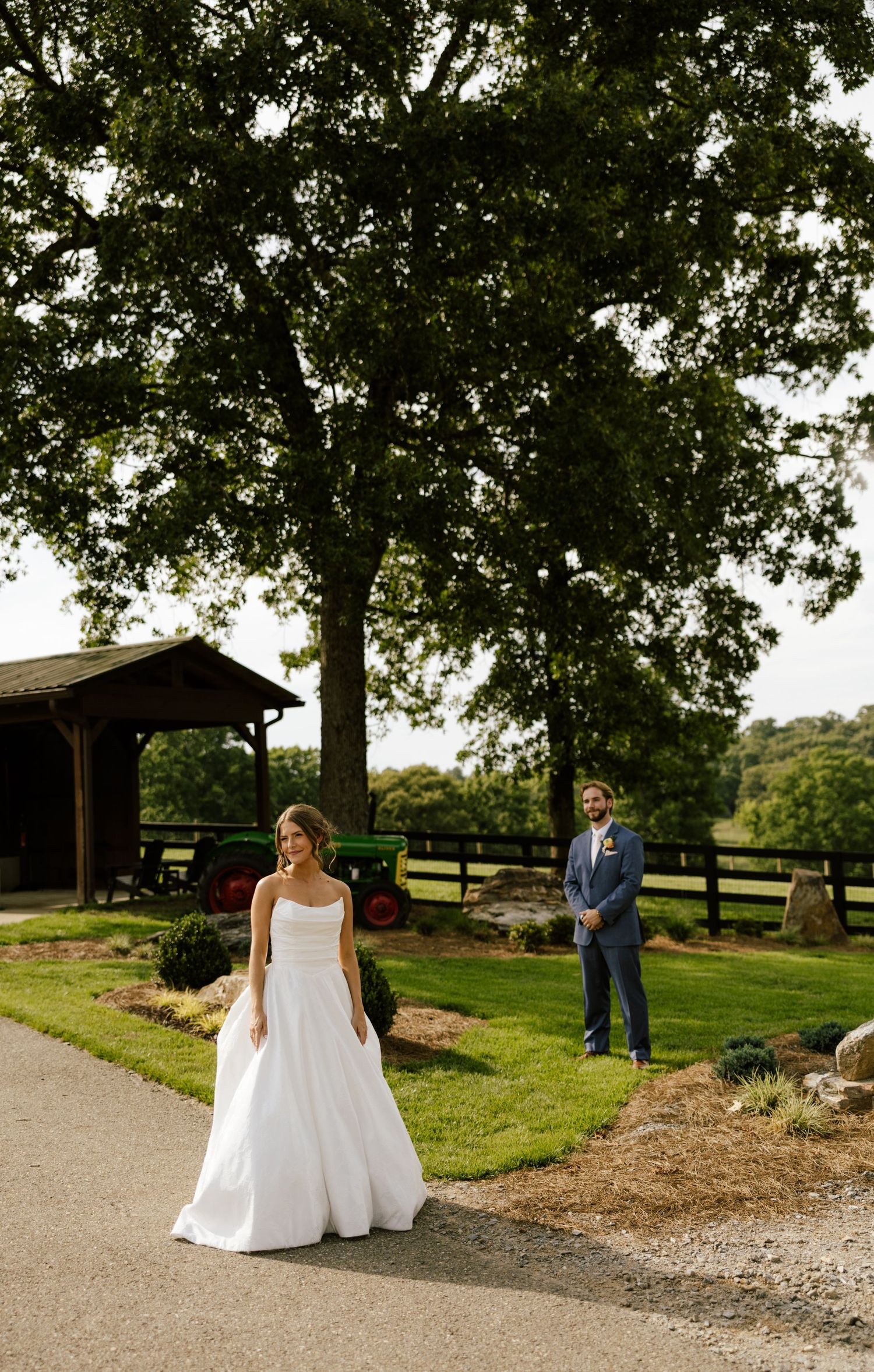 Bride in white gown and groom in blue suit share a first look moment on a rustic farm path with green trees.