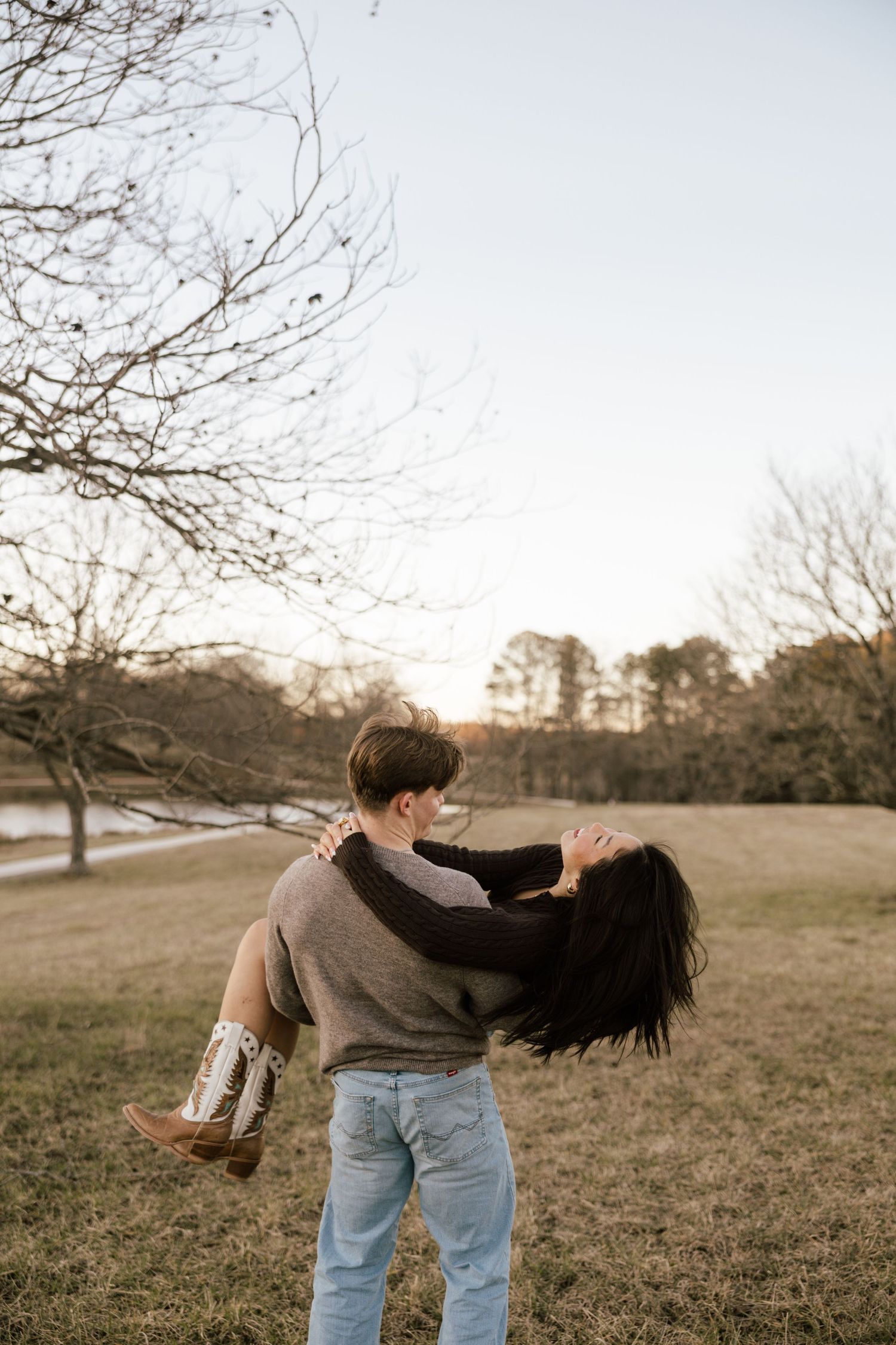 A man lifts a woman in his arms outdoors in a park during golden hour, surrounded by bare trees.