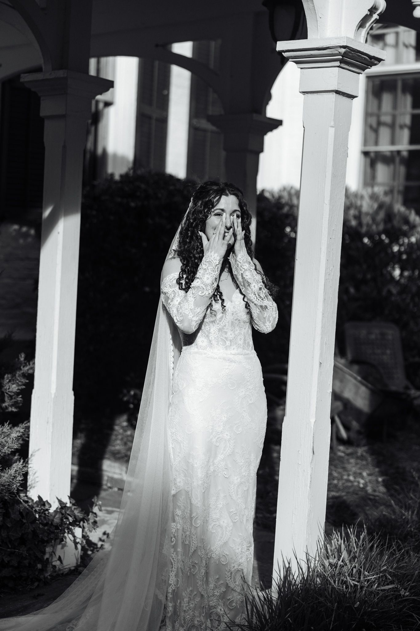 Black and white photo of an emotional bride in lace gown and veil covering her mouth with hands near white columns.