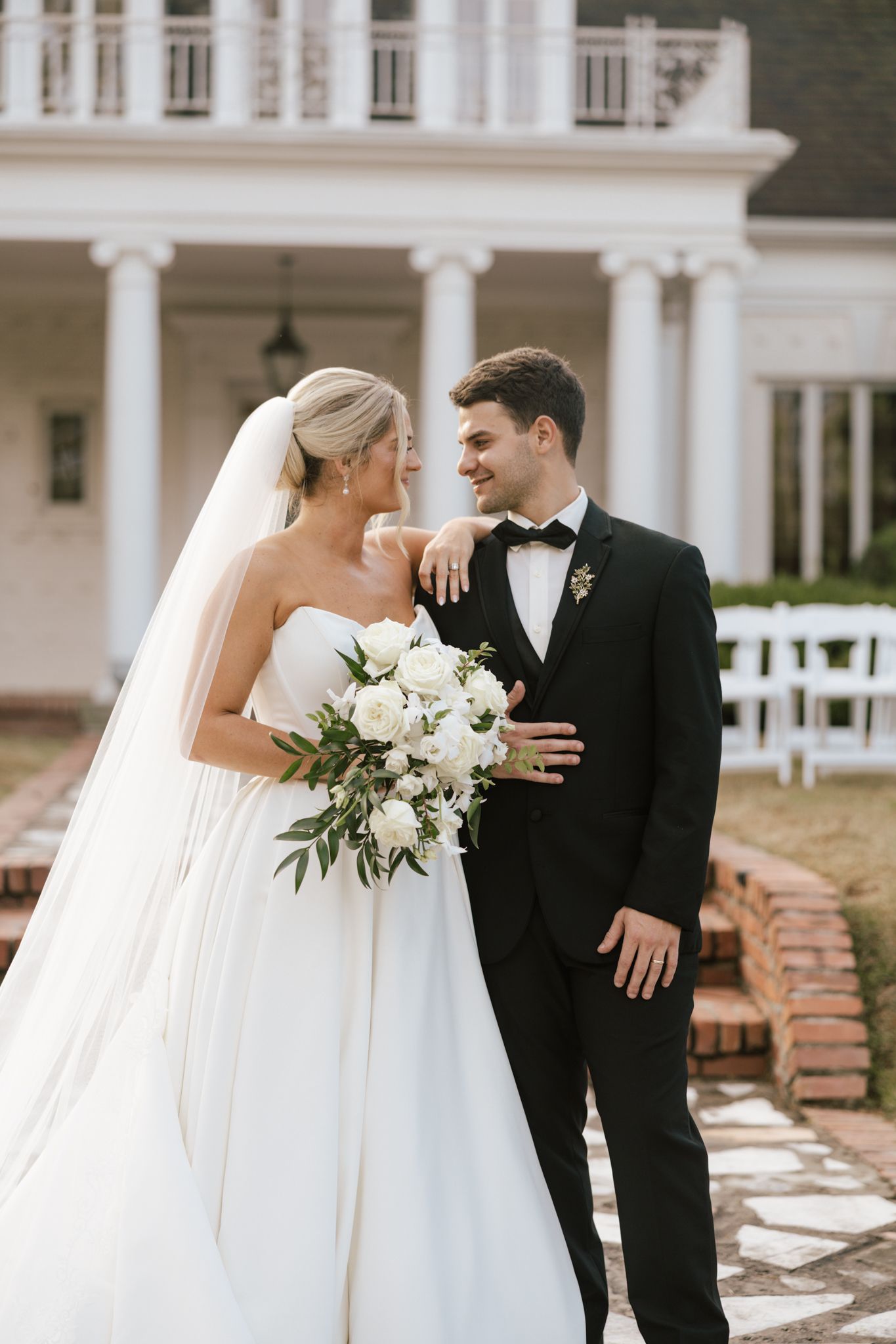 Bride in strapless white ball gown with veil holding white bouquet, groom in black tuxedo, smiling outside elegant white mansion.
