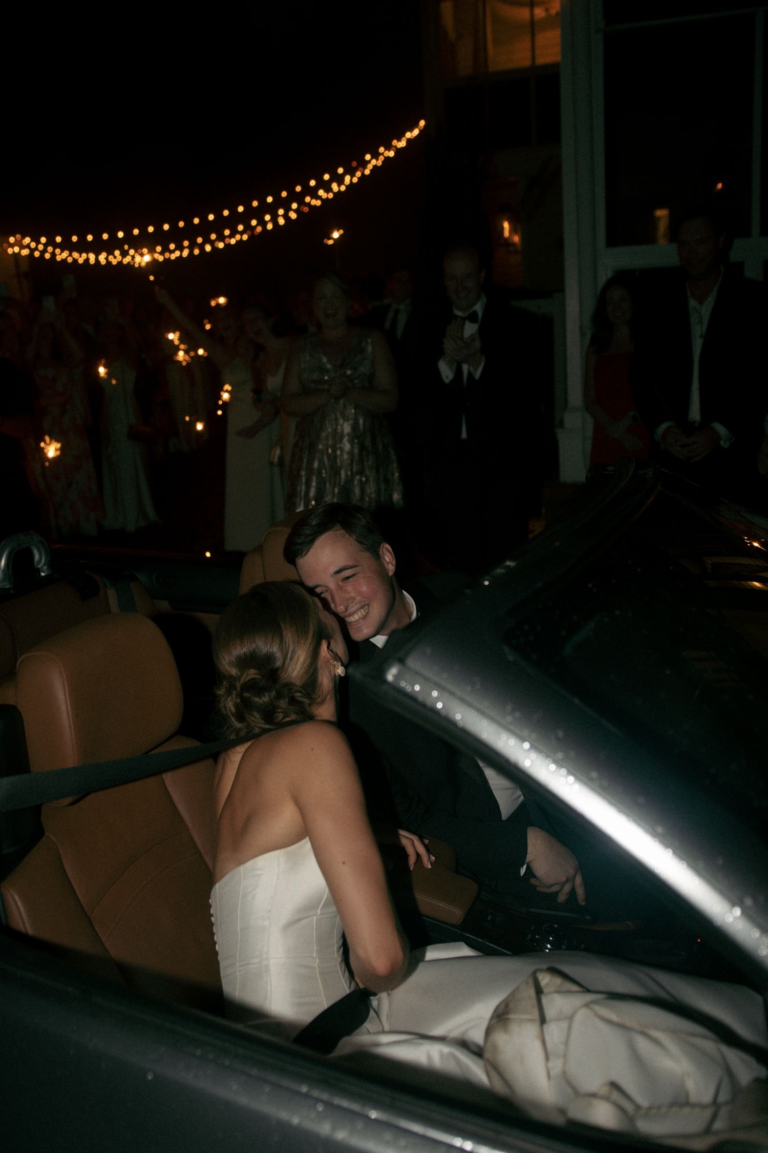 Bride and groom sharing a romantic moment in a convertible car at night, surrounded by wedding guests holding sparklers.