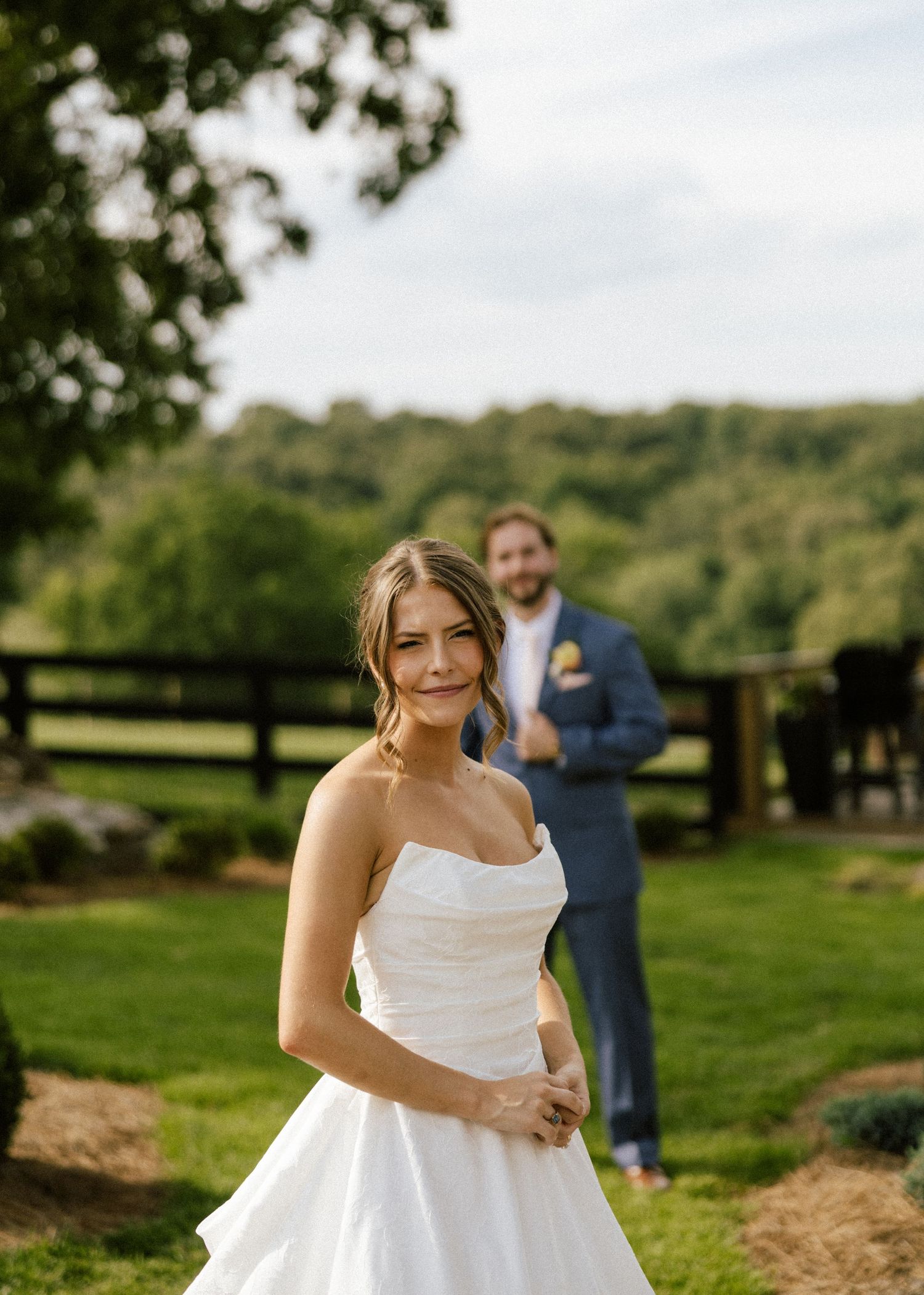Bride in strapless white gown smiles outdoors with groom in blue suit standing in background at rustic farm venue.