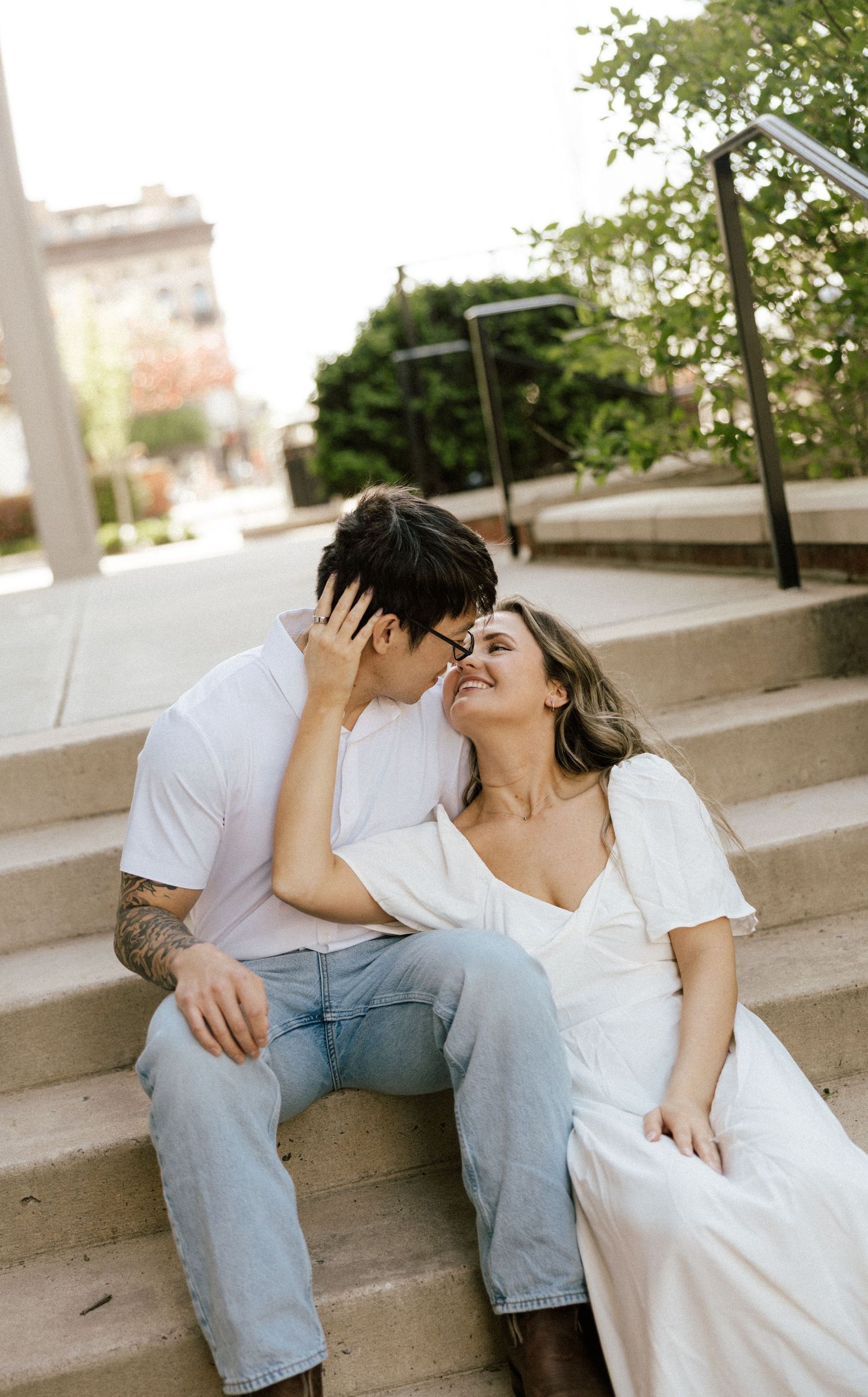 A couple sits on outdoor steps, sharing a romantic almost-kiss, dressed in white tops and light jeans.