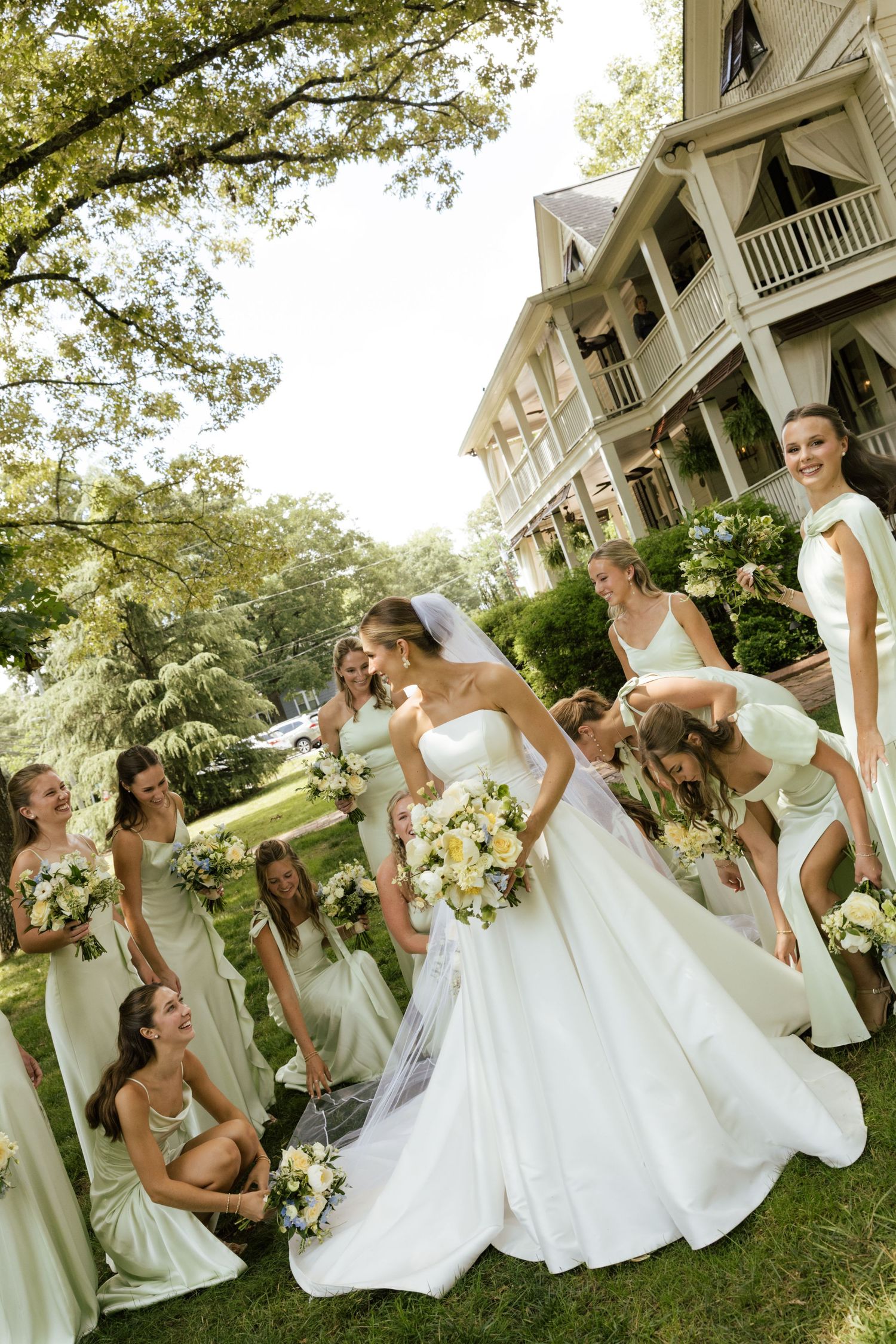 Bride in white gown surrounded by bridesmaids in sage green dresses holding bouquets on lawn outside historic mansion.