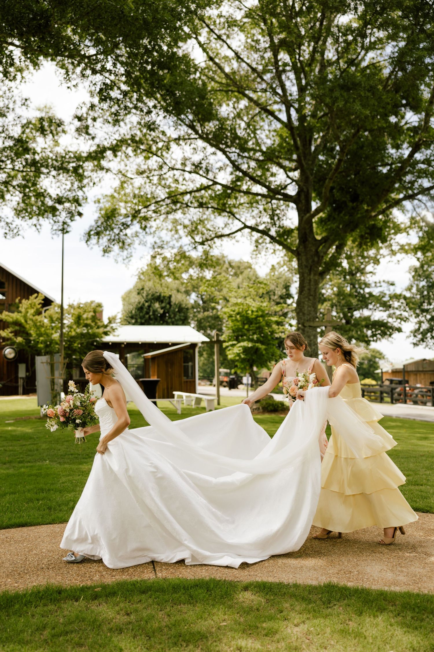 Bride in white gown with two bridesmaids in yellow dresses arranging her train outdoors near large trees.