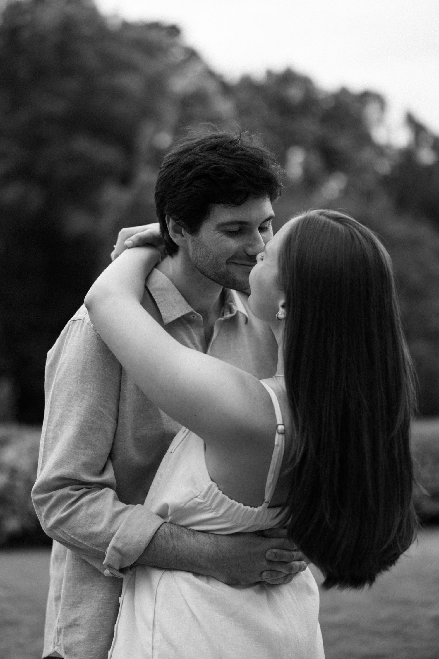 Black and white photo of a couple embracing and kissing outdoors with trees in the background.
