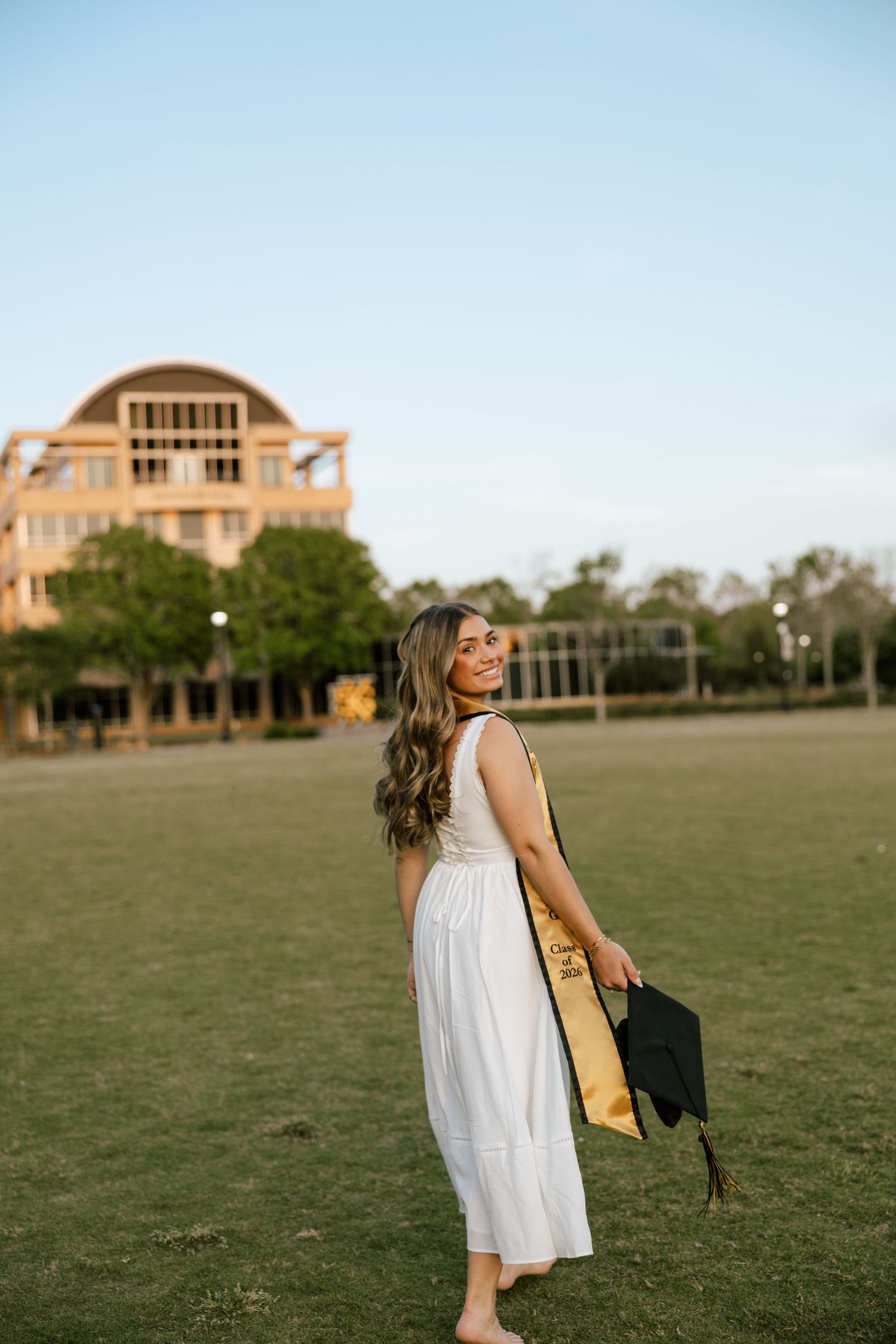 Young woman in white dress holding graduation cap and gold stole, smiling on green campus lawn at golden hour.