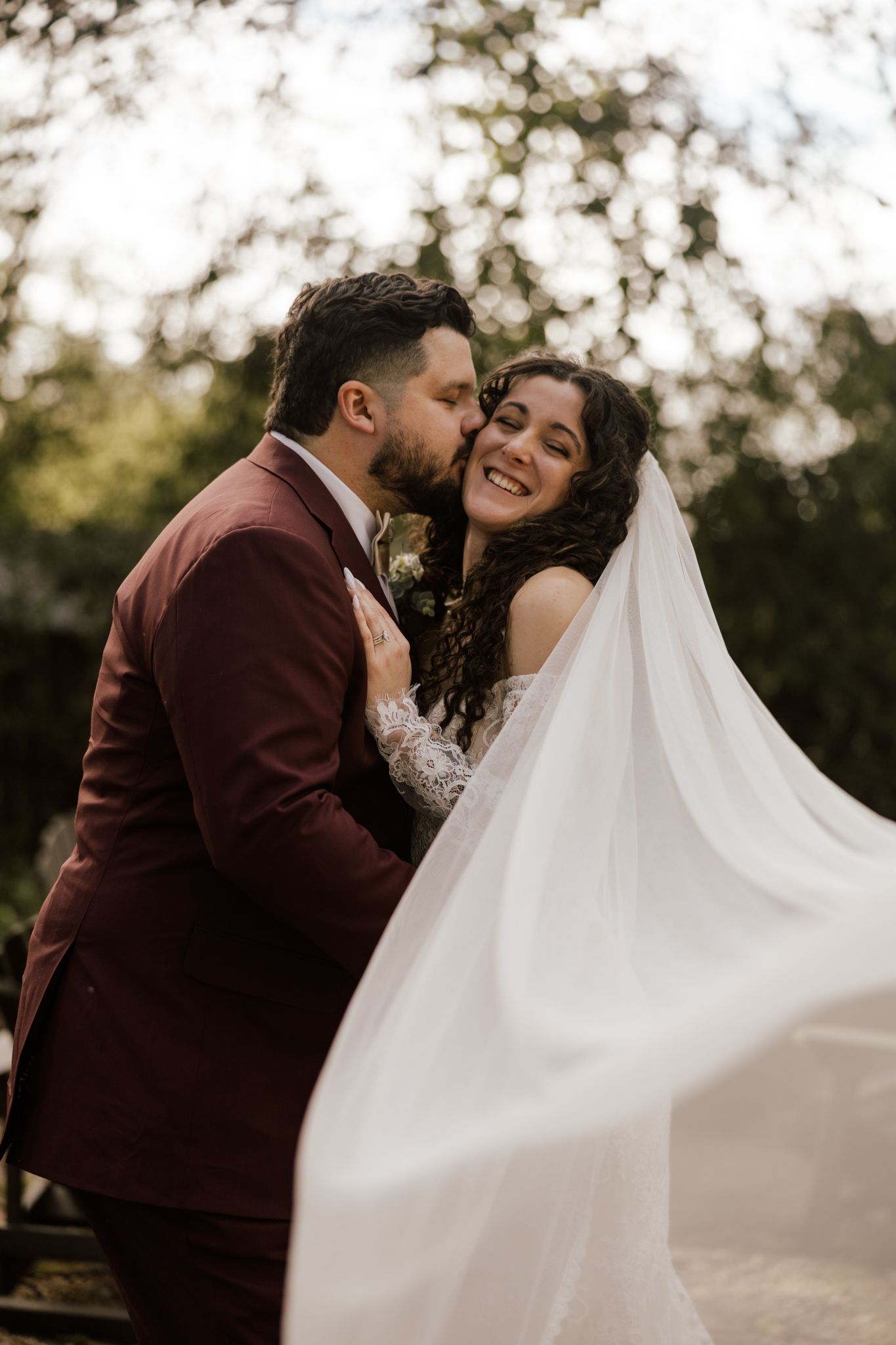 Groom in burgundy suit kisses bride on cheek as her veil flows dramatically in the breeze.