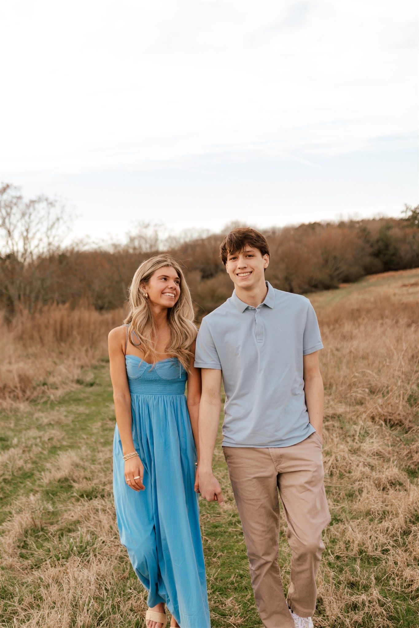 Couple in blue dress and polo shirt walking together through golden field at sunset.