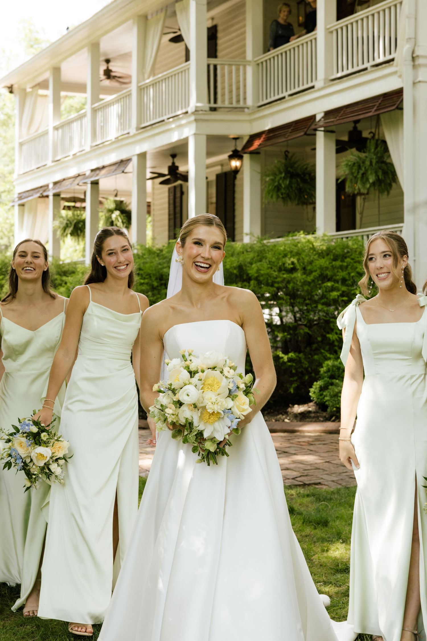 Bride in white strapless gown walks with bridesmaids in sage dresses, holding bouquets outside elegant venue.