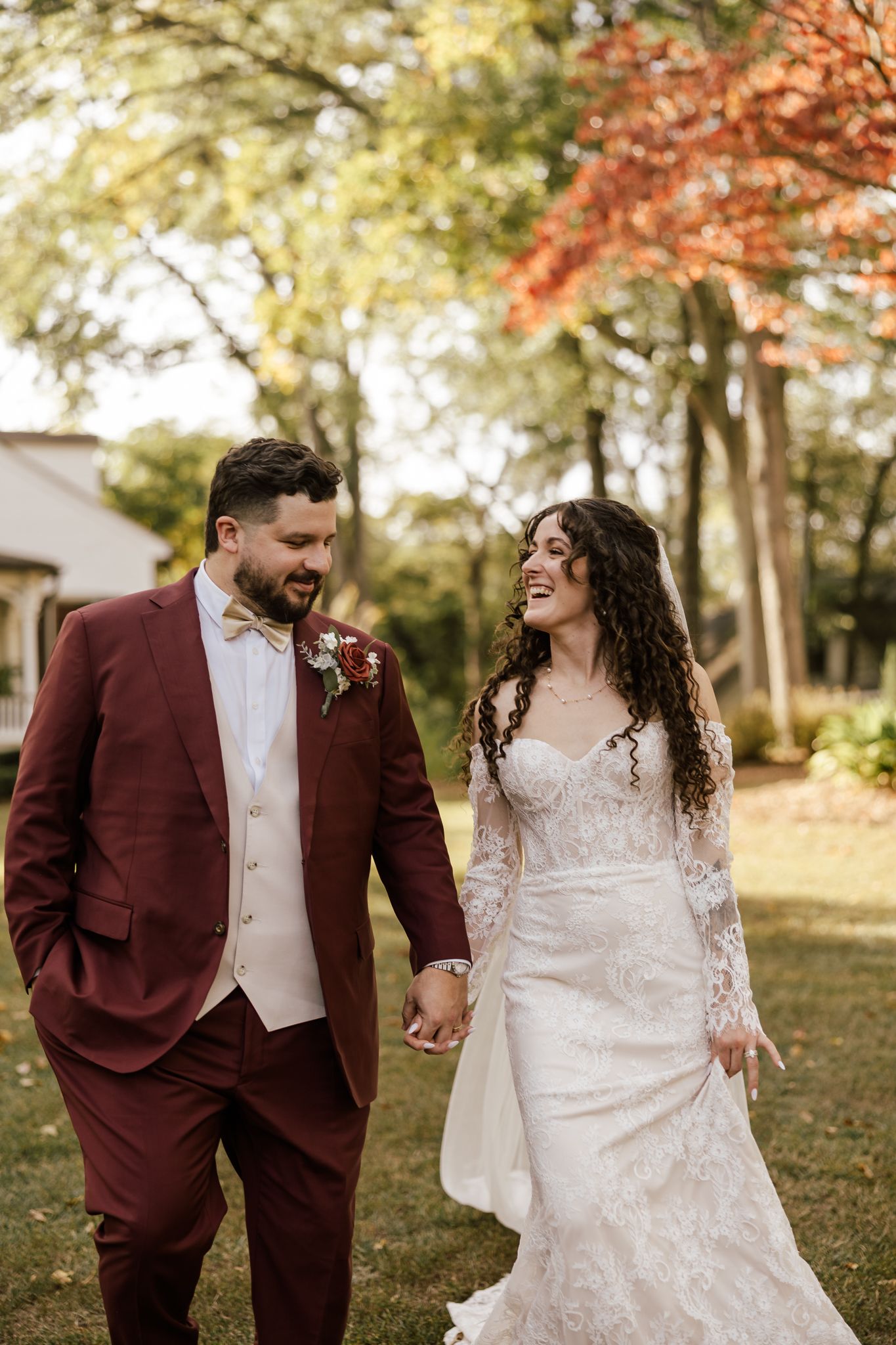Bride and groom holding hands walking outdoors, groom in burgundy suit, bride in lace wedding dress.