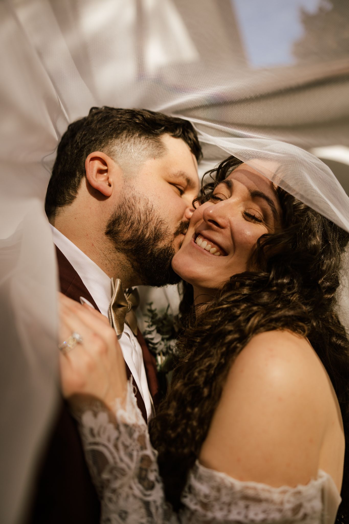 Groom kisses smiling bride on the cheek under a flowing veil during romantic wedding ceremony moment.