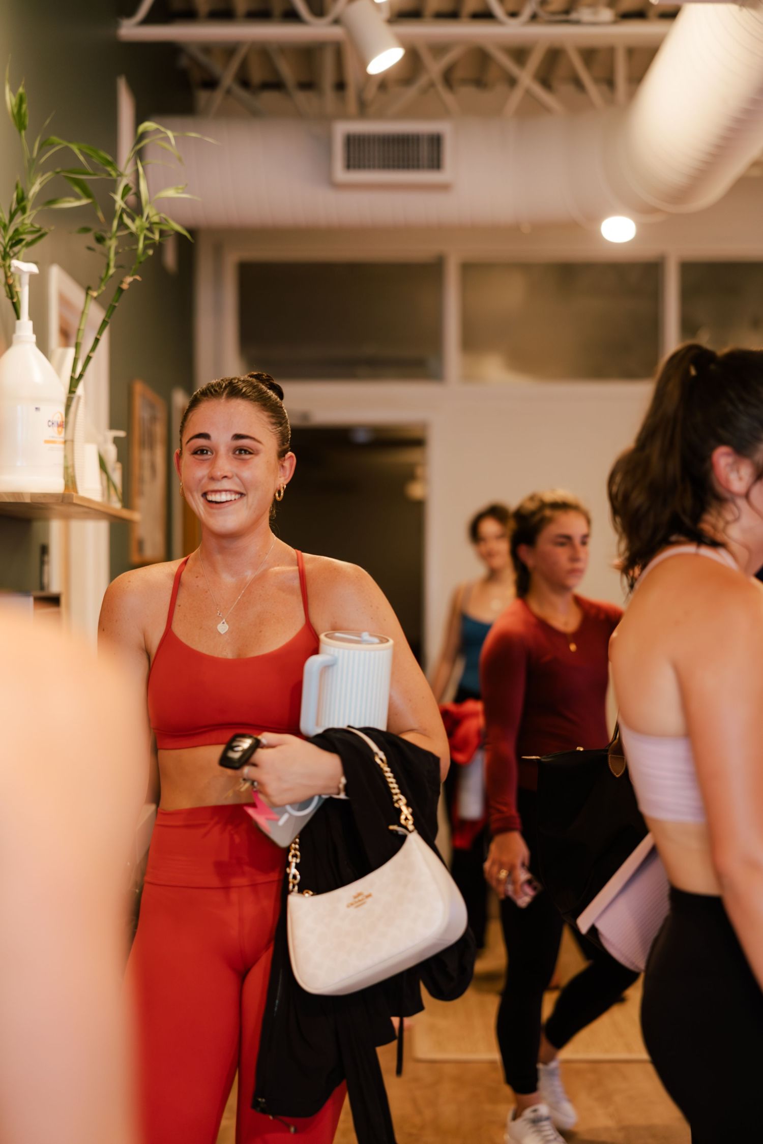 Smiling woman in red athletic wear holding a water bottle and white purse at a yoga studio.
