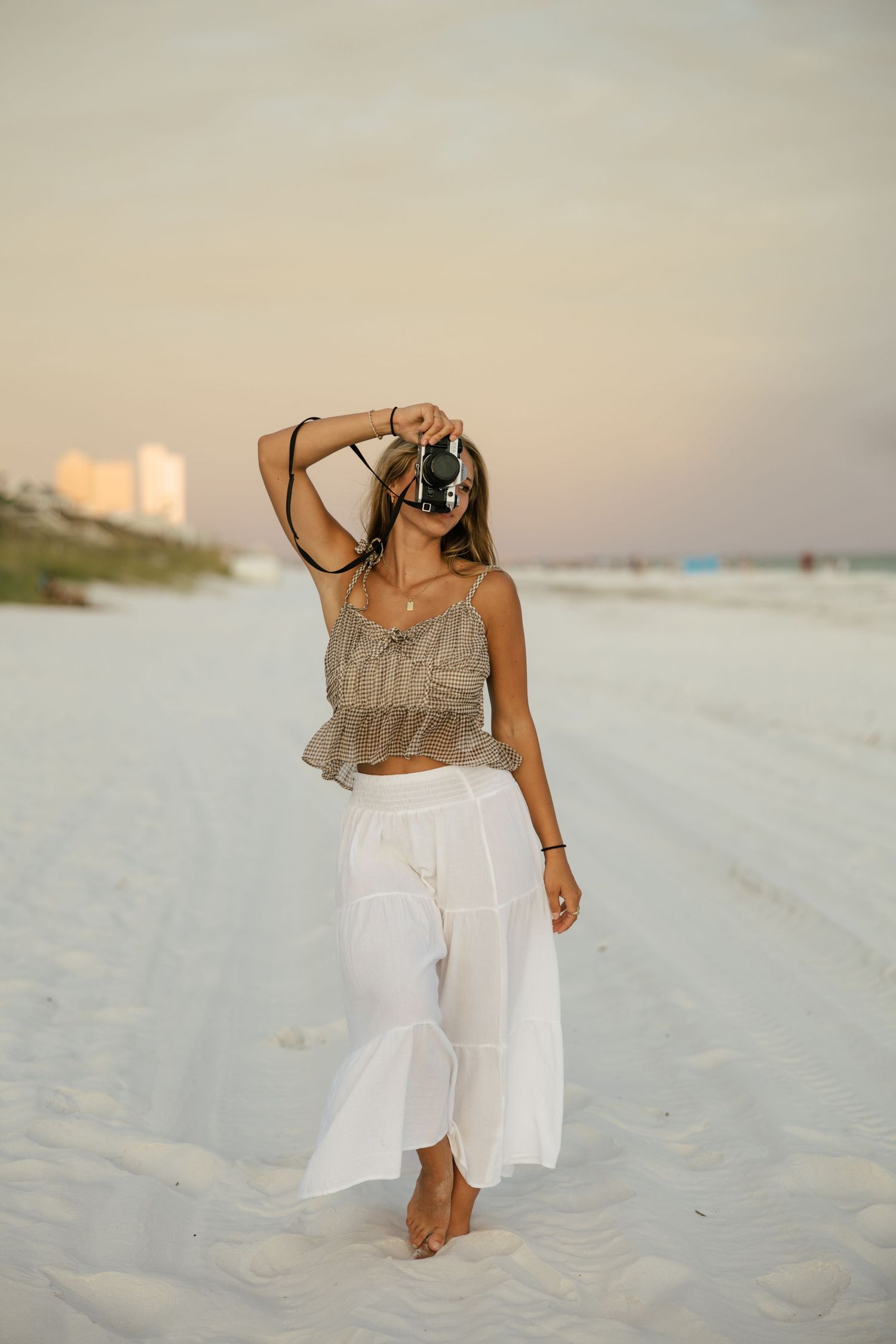 Woman in white pants and crop top holds camera on white sand beach at sunset.