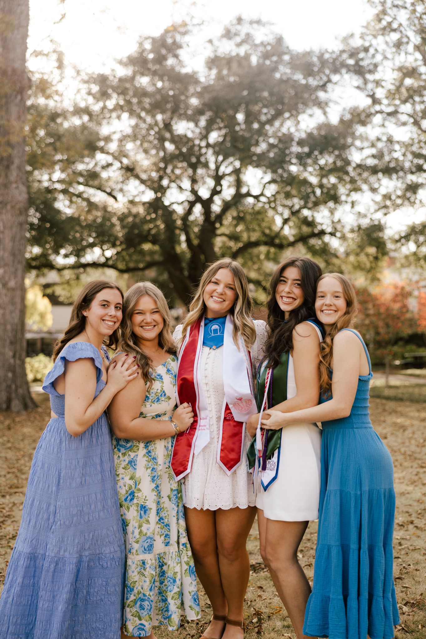 Five young women pose outdoors at a graduation celebration, one wearing a white dress with colorful academic stoles.