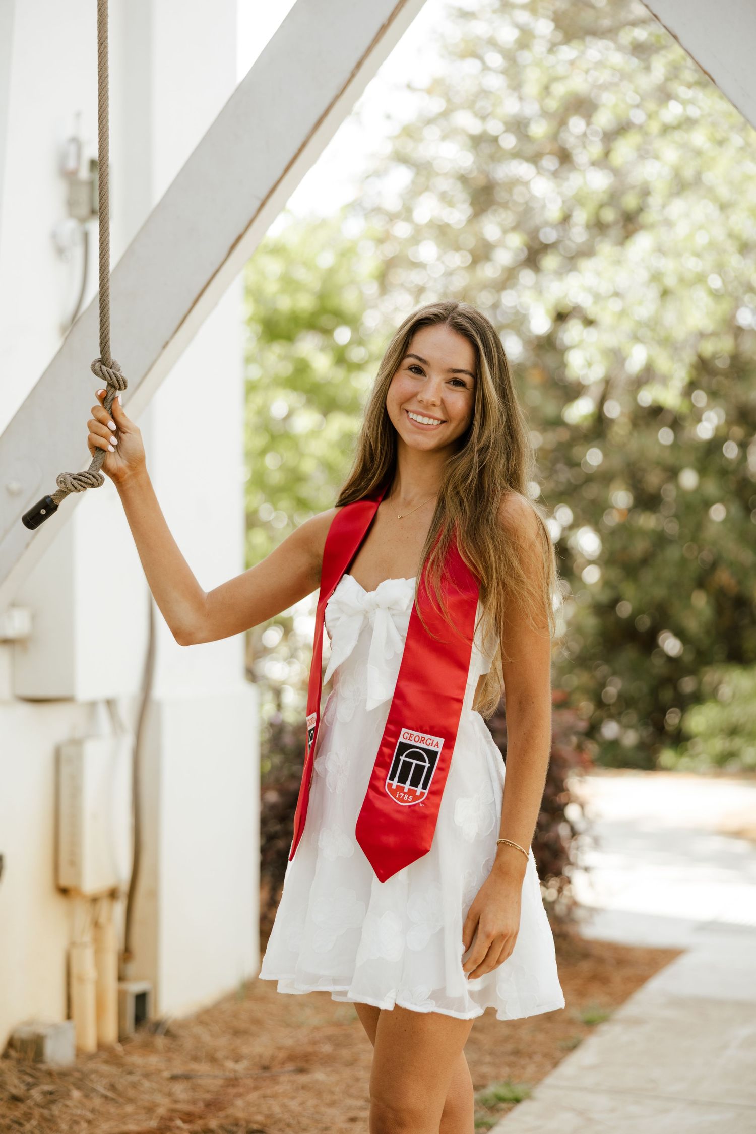 Smiling young woman in white dress and red graduation stole posing outdoors for senior photos.