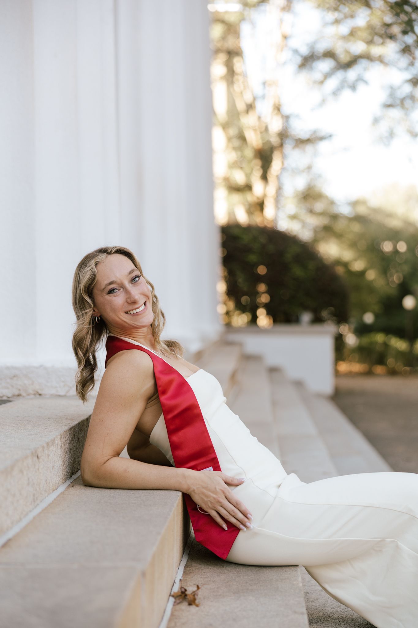Graduate in red and white outfit sits casually on white stone steps during outdoor photoshoot.