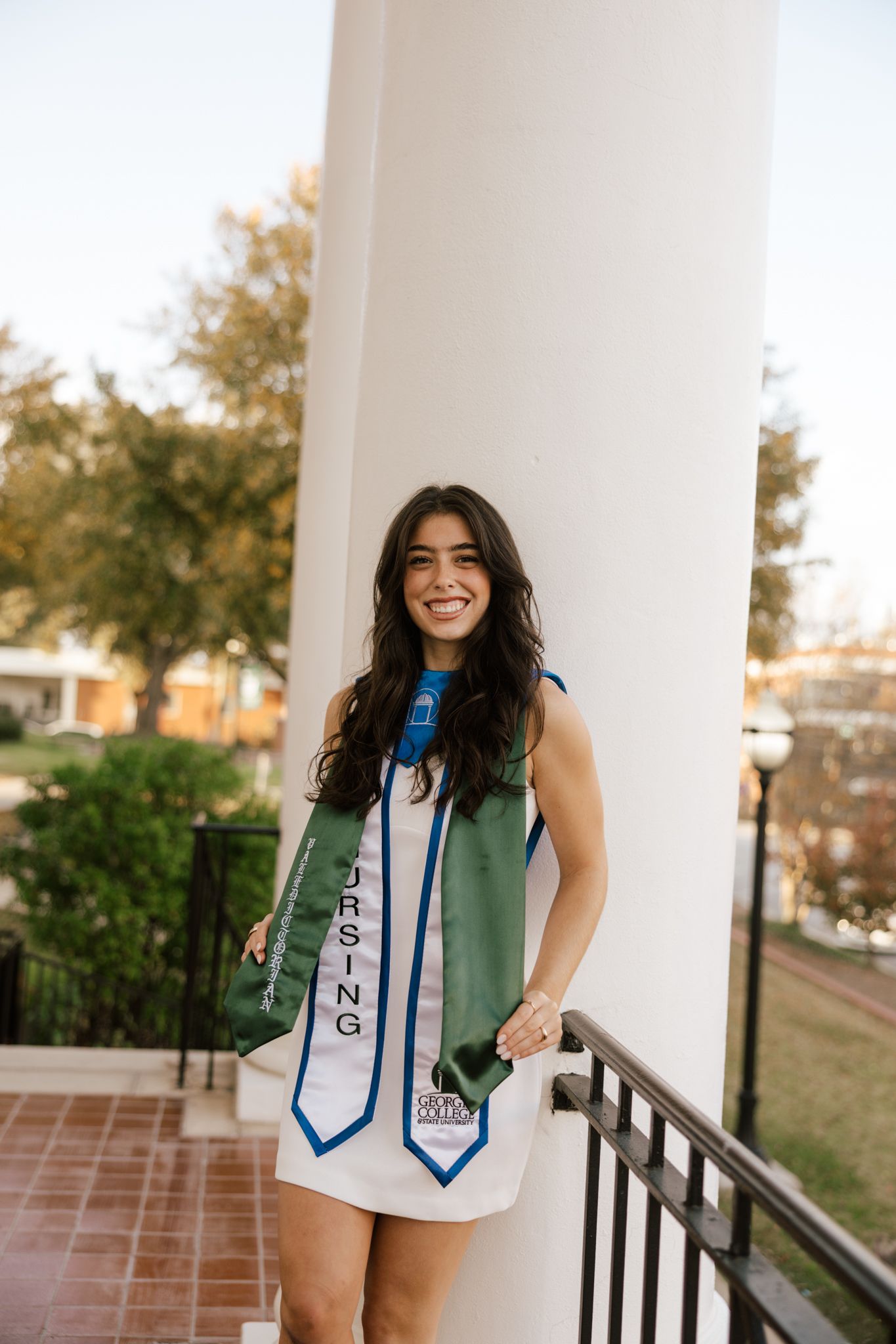 Graduate in white dress with decorative sashes posing by white columns outdoors.