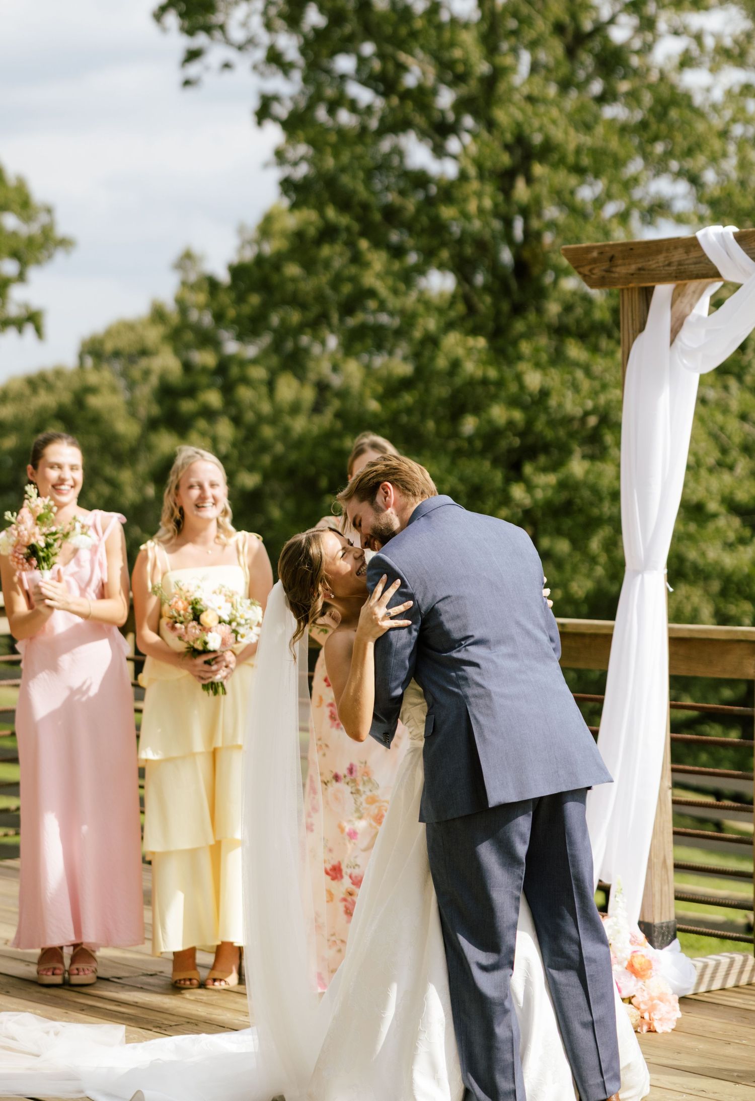 Bride and groom share first kiss at outdoor wedding ceremony while bridesmaids in pink and yellow dresses look on.