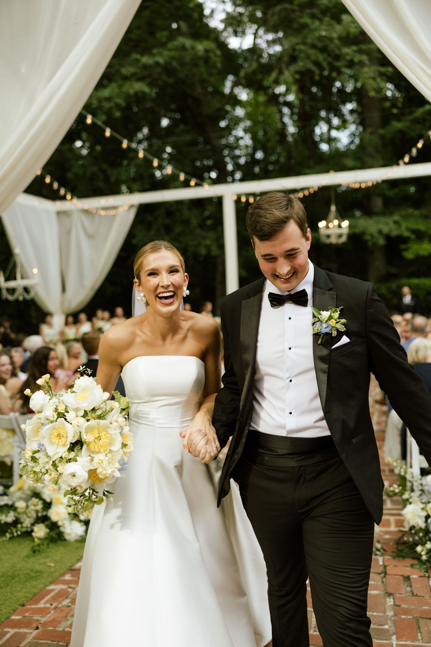 Joyful bride in strapless white gown holding floral bouquet walks with groom in black tuxedo after outdoor wedding ceremony.