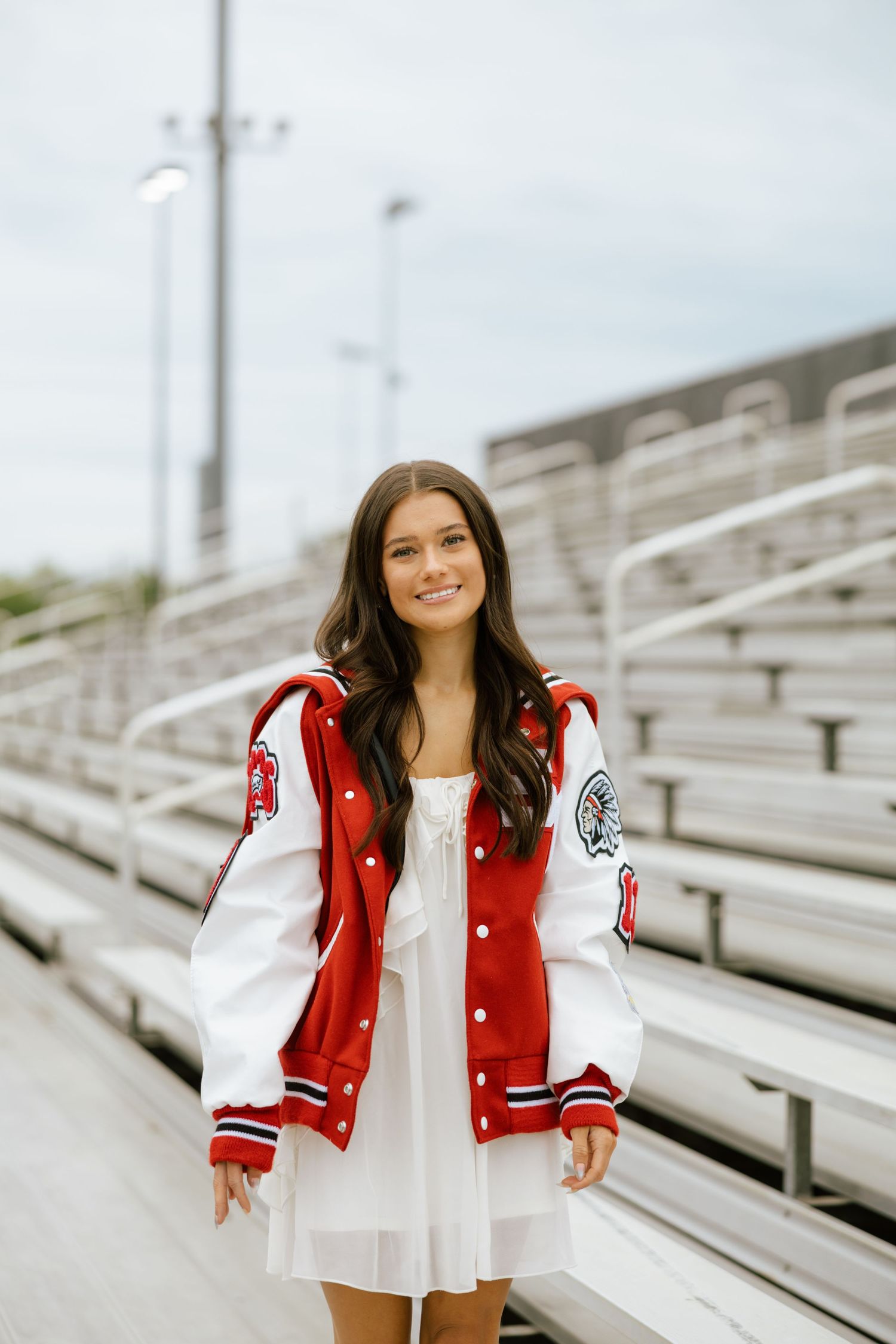 Young woman in red and white varsity jacket over white dress smiling in bleachers at a high school stadium.