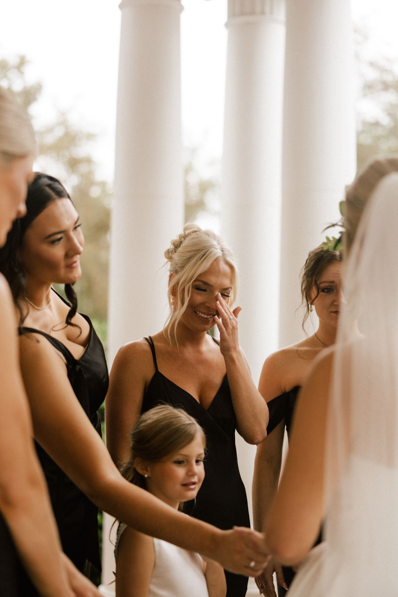 Bridesmaid wiping tears while holding hands with bride surrounded by bridesmaids in black dresses near white columns.