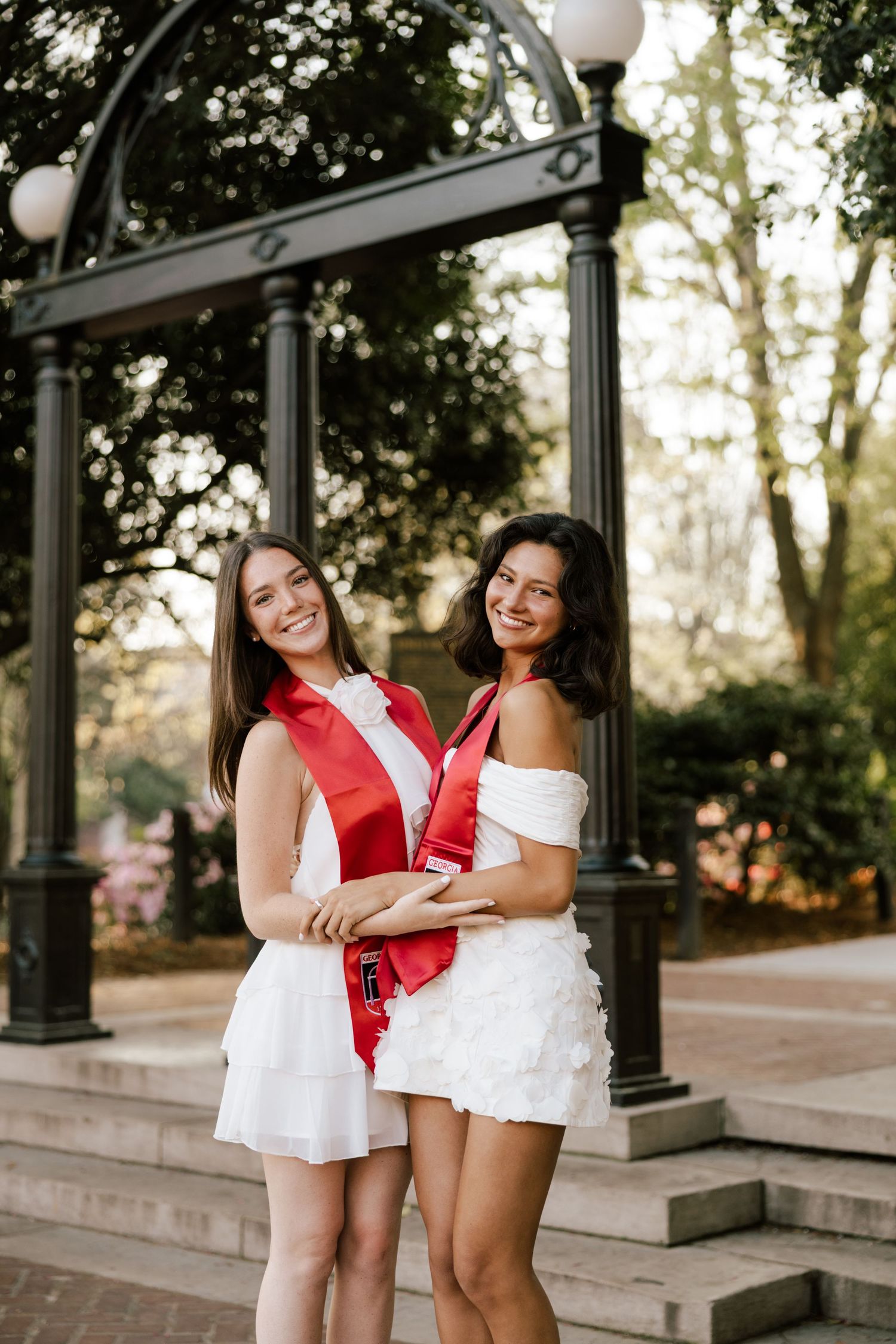 Two smiling women in white dresses and red graduation stoles pose outdoors near a decorative arch.