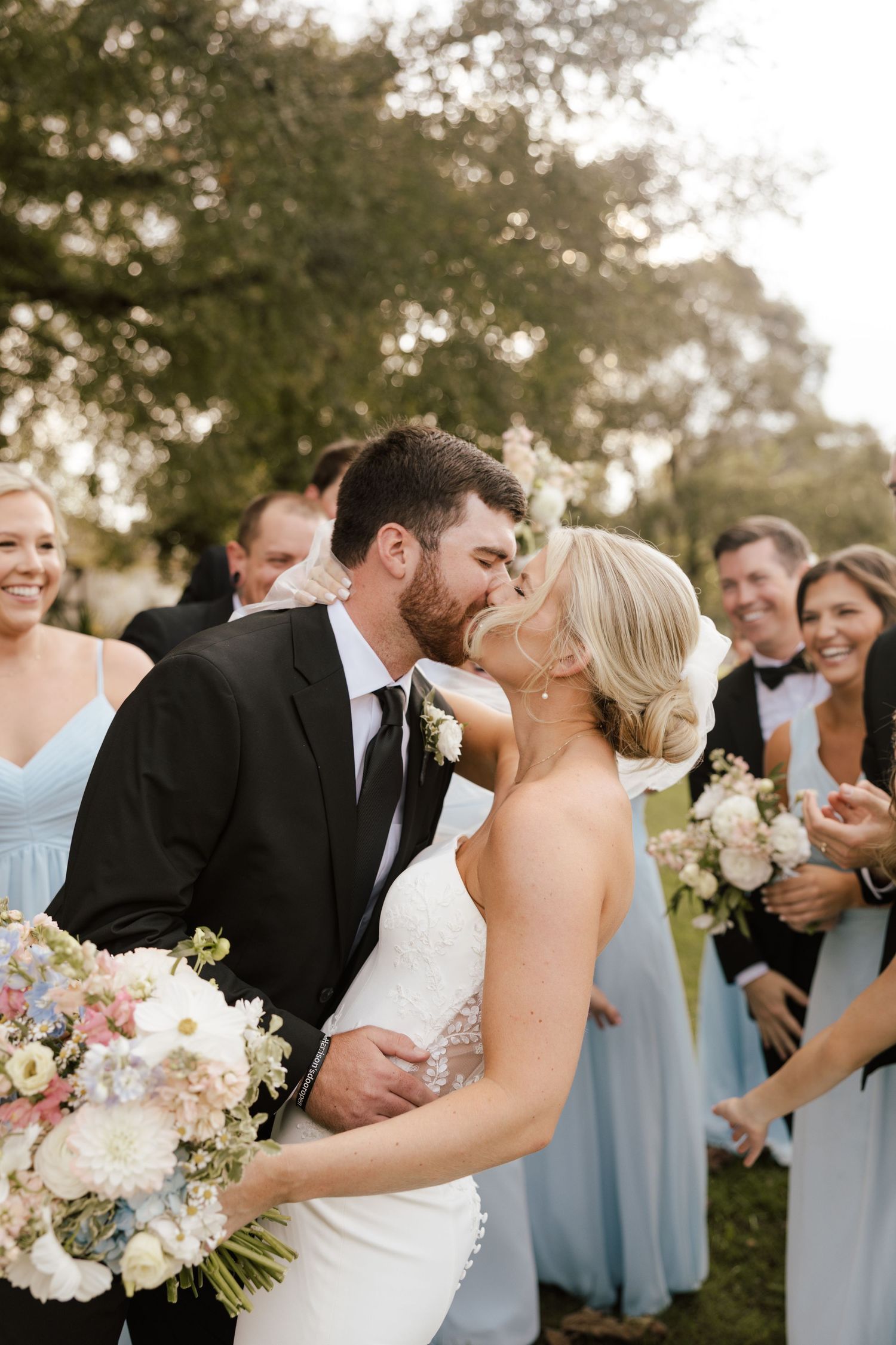 Newlyweds share a first kiss surrounded by smiling bridesmaids in light blue dresses and groomsmen.