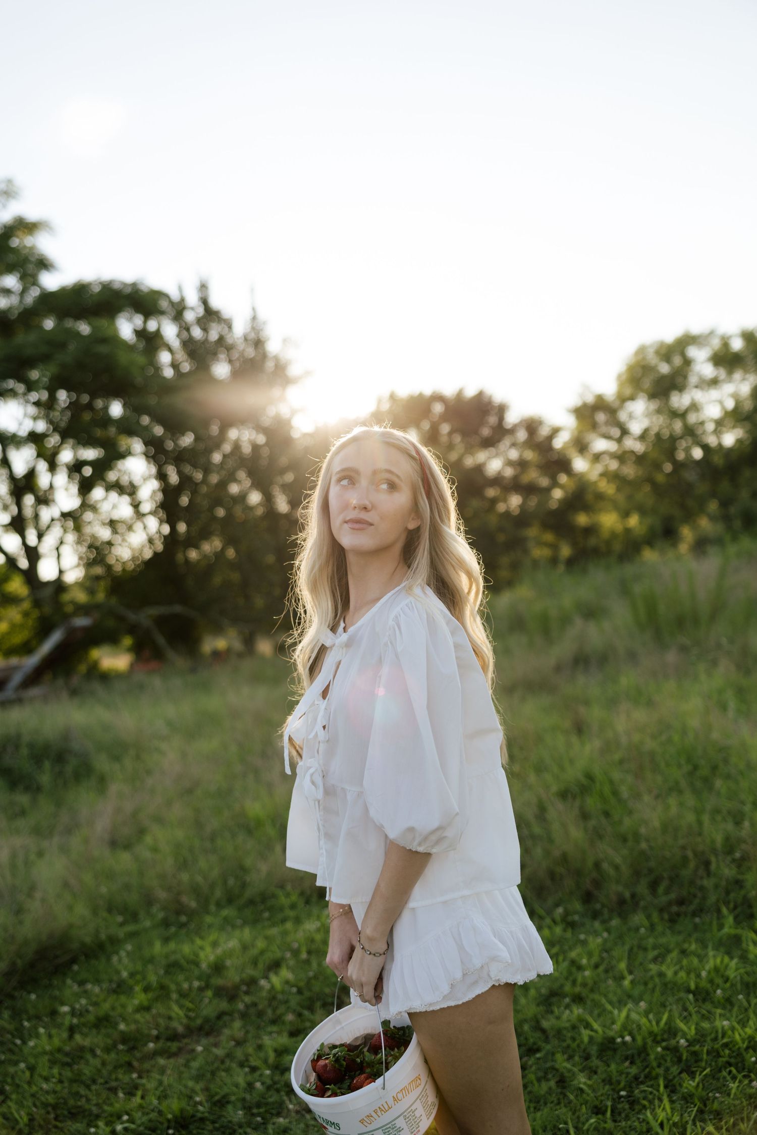 Young woman in white dress holding a bucket of cherries in a sunlit green field at golden hour.