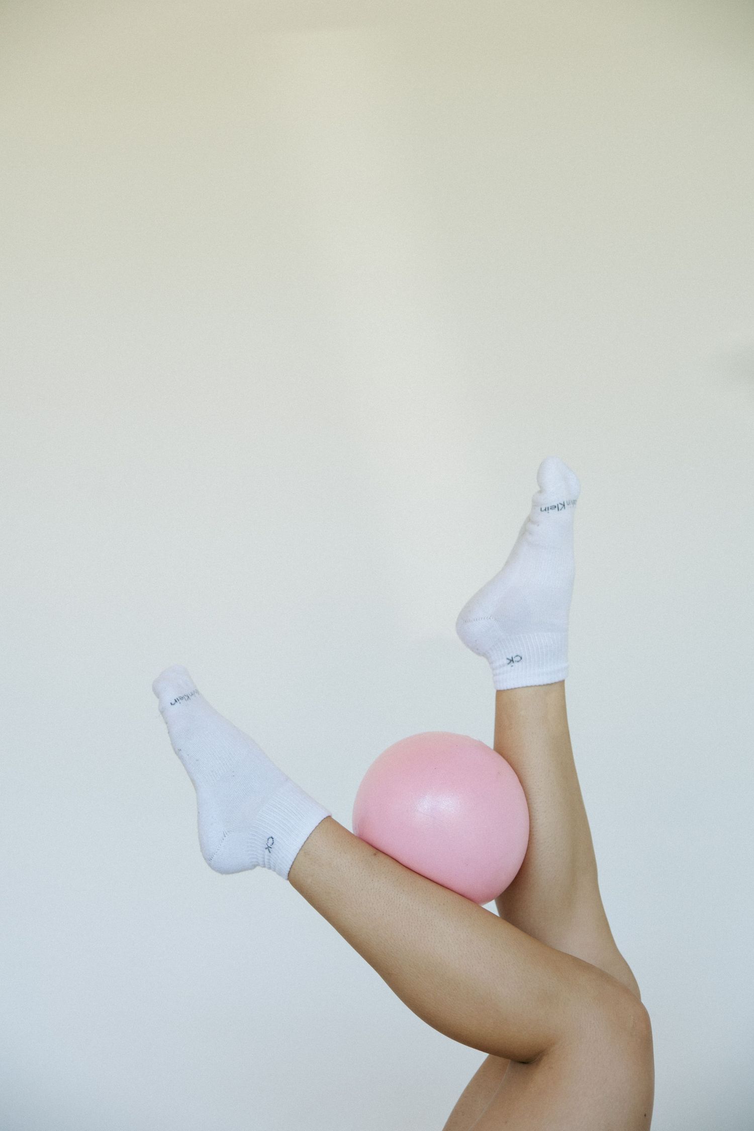 Legs raised in air wearing white socks with pink exercise ball balanced between feet against white background.