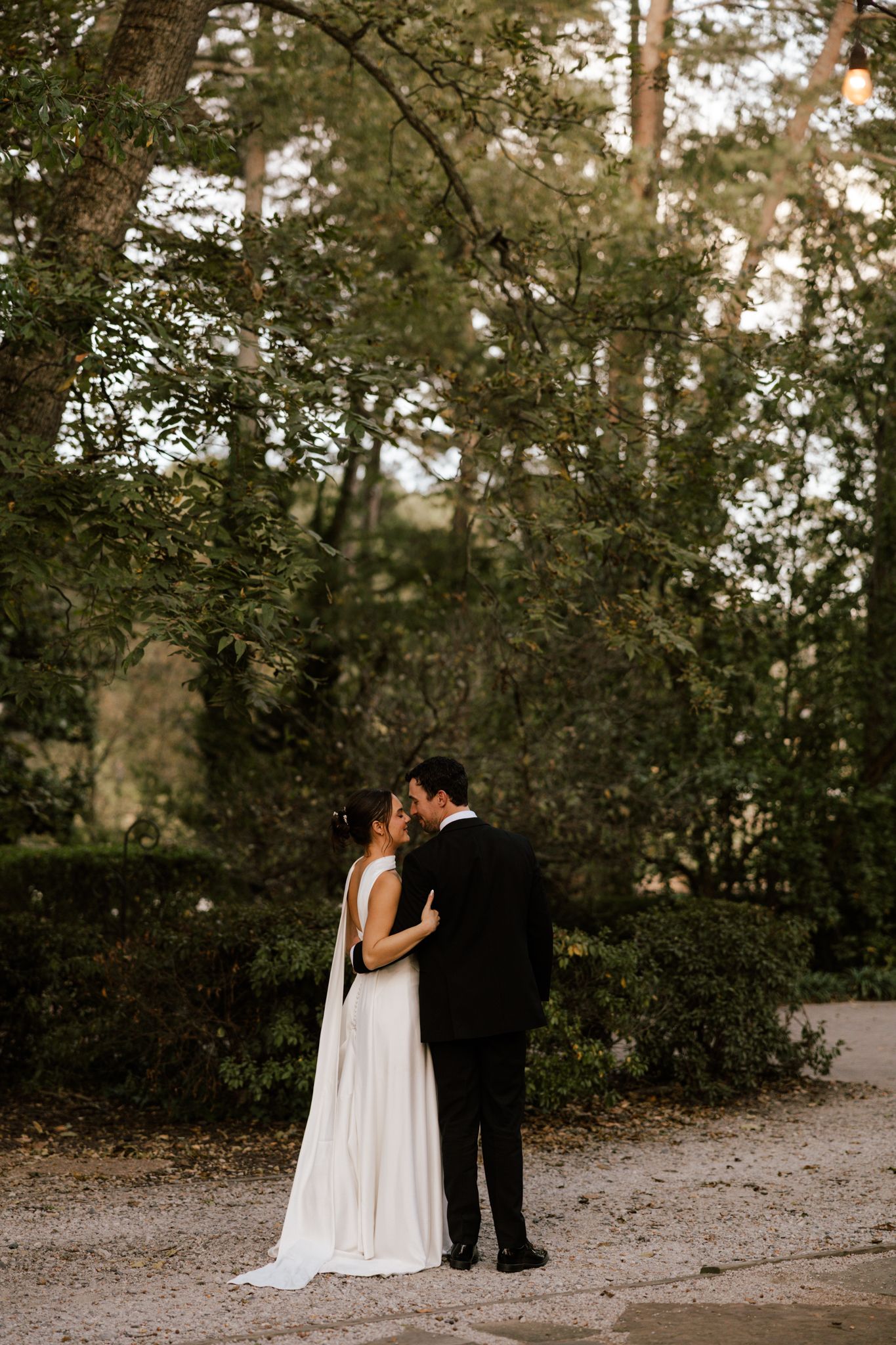 Bride and groom share a romantic kiss outdoors surrounded by lush green trees in a serene garden setting.