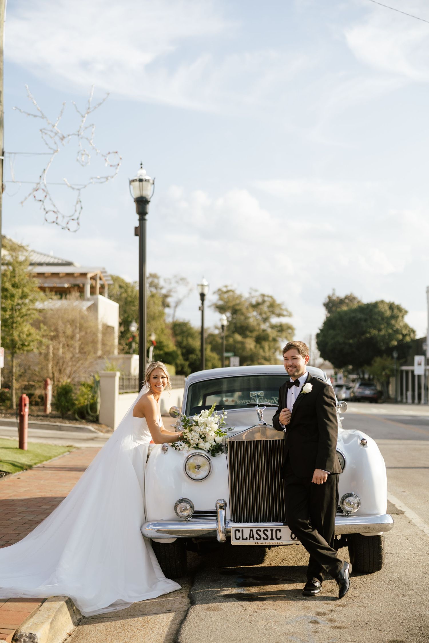 Bride and groom posing beside a classic white vintage car on their wedding day on a sunny street.