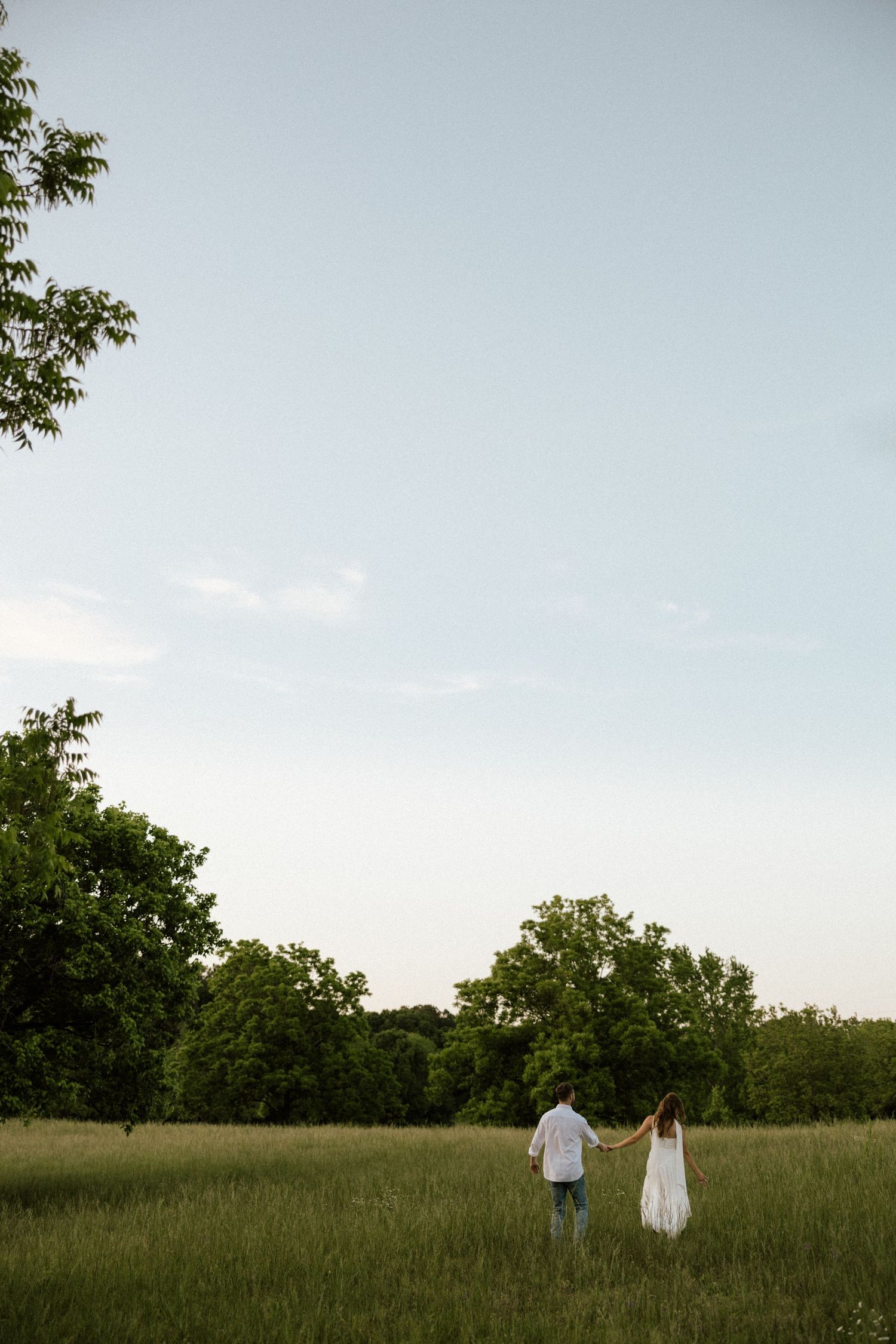 Couple holding hands walking through a lush green meadow with trees in the background under a soft blue sky.